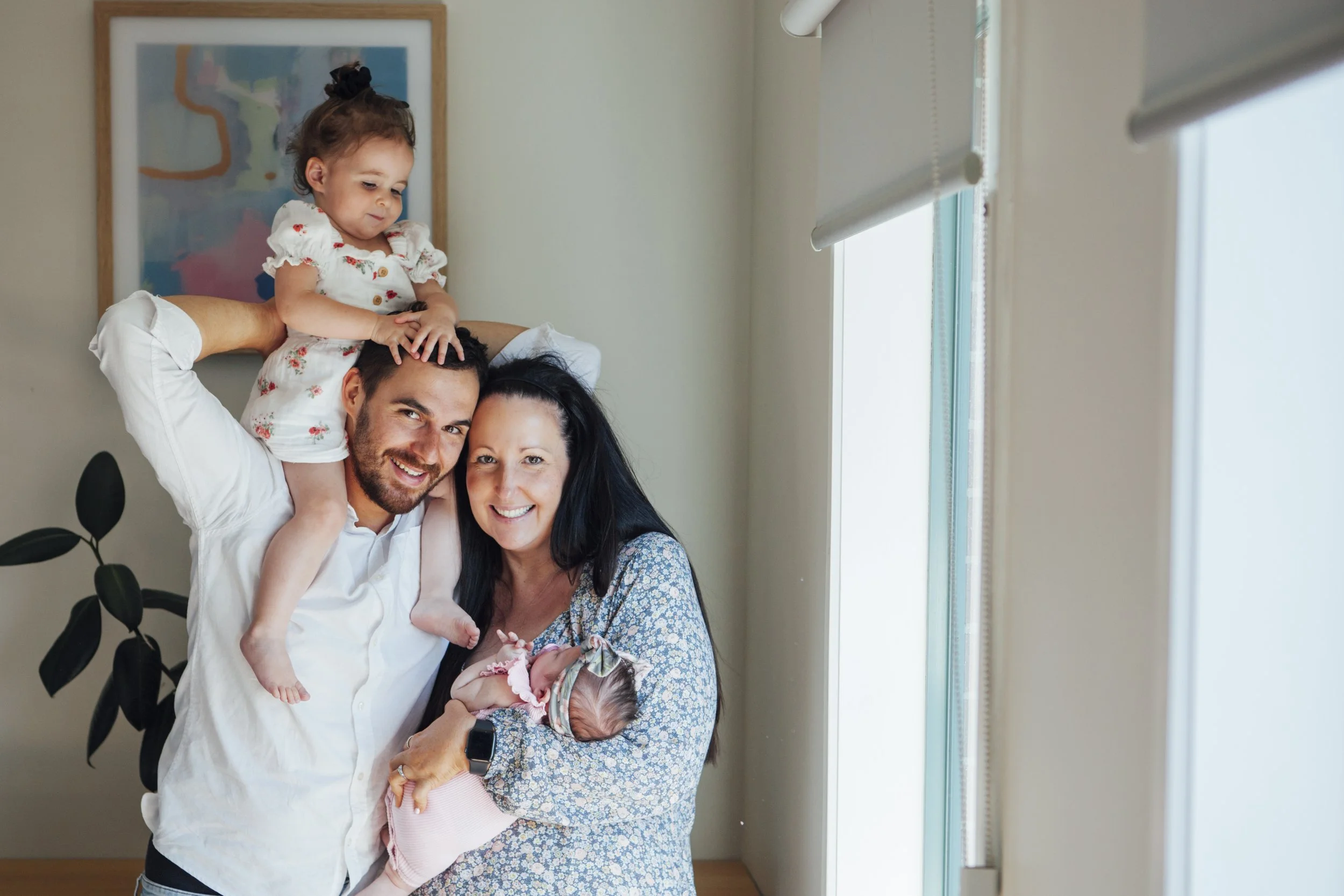 A family of four, including a mother, father, young girl, and newborn baby, celebrating indoors with natural light.