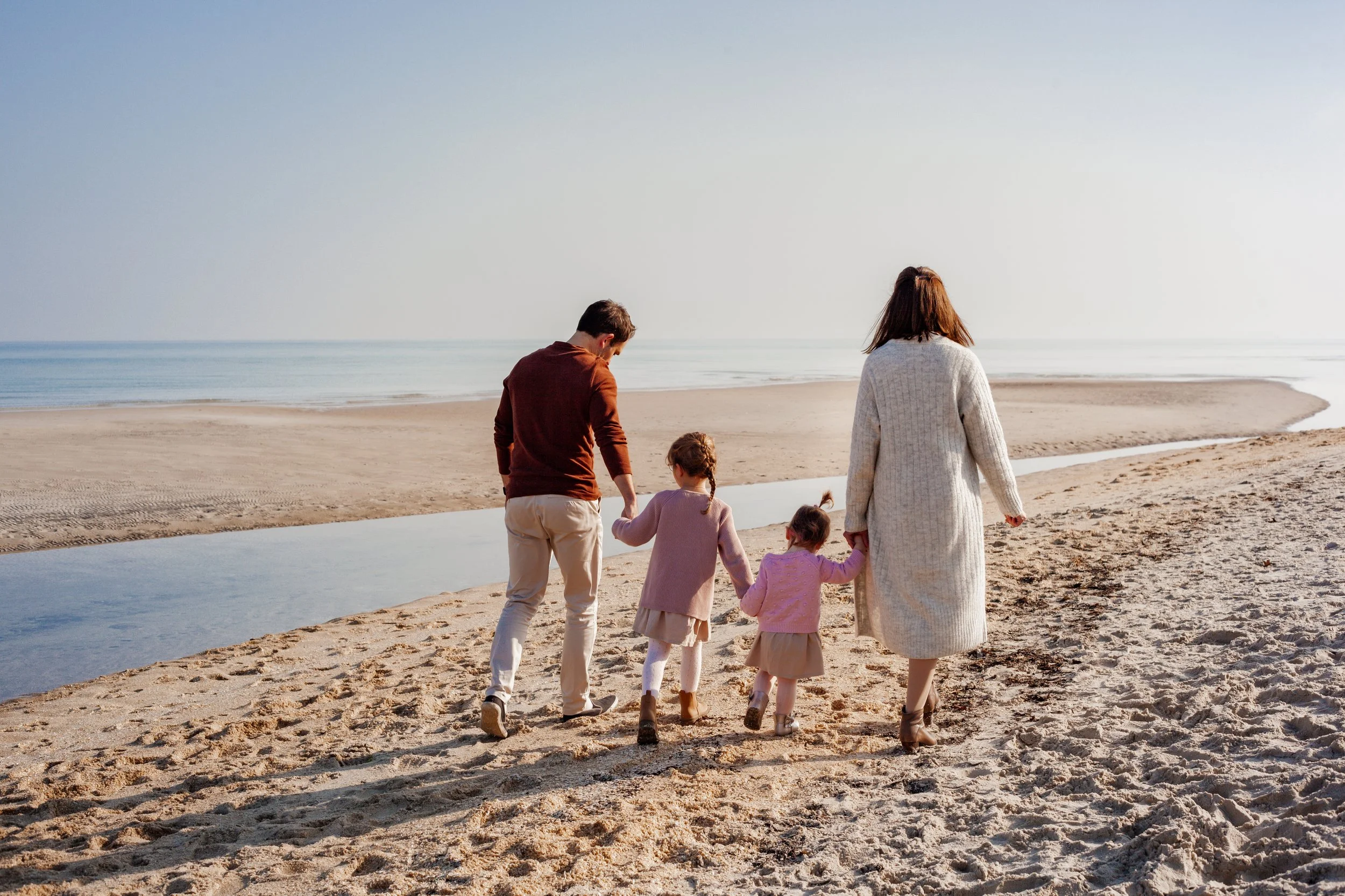 Family of four walking on a sandy beach near the ocean, with a dad, mom, and two young girls holding hands.