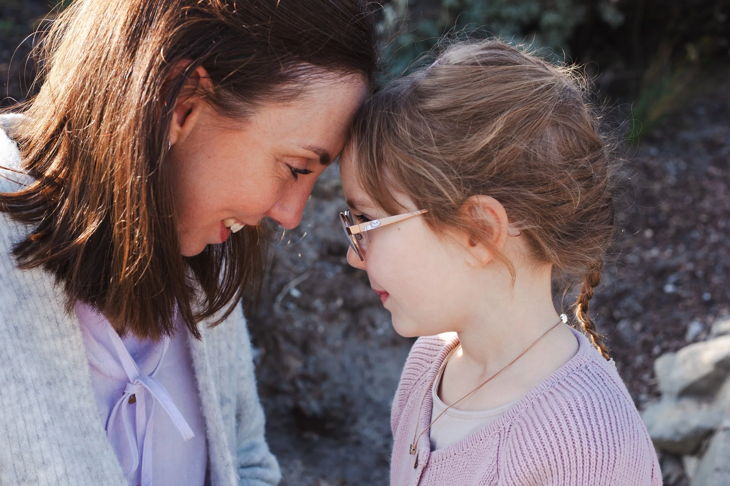 A woman and a young girl with their foreheads touching and eyes closed, outdoors, sharing a tender moment.
