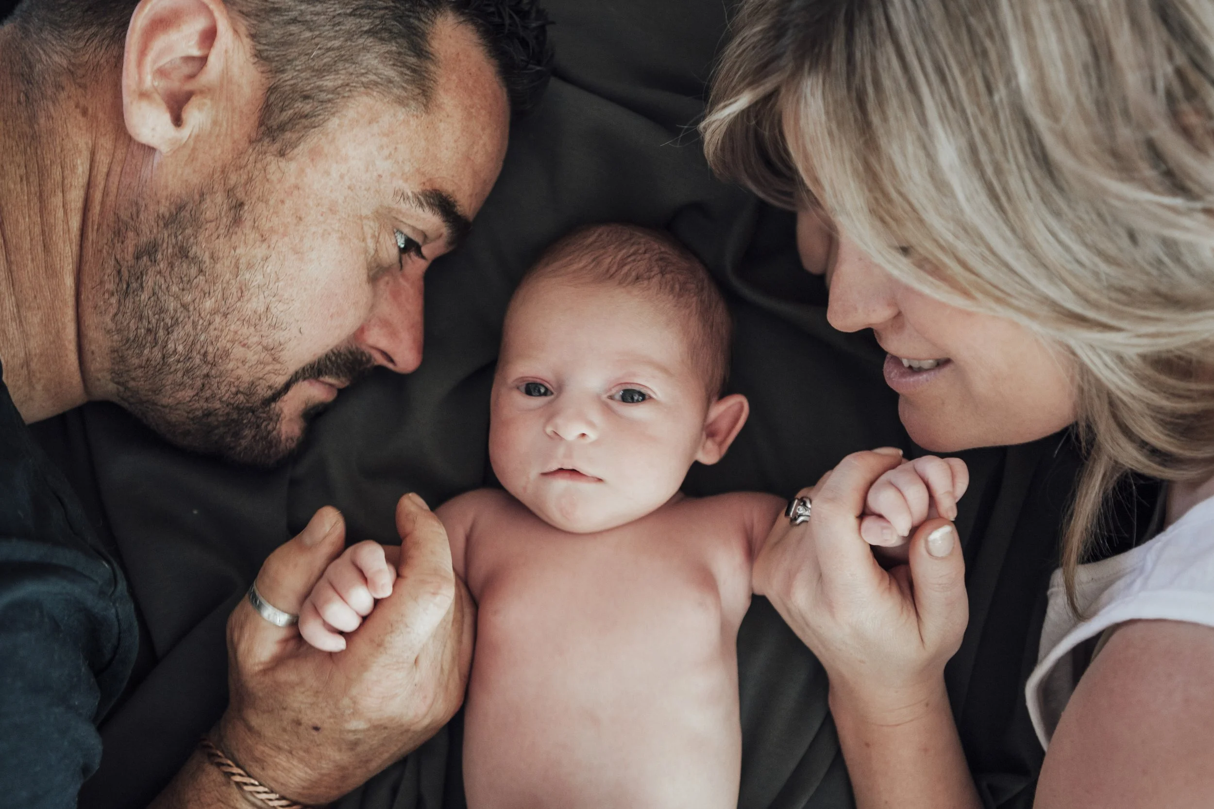 A photo of a baby lying between a man and a woman with their foreheads touching, all looking at the camera. The baby is shirtless, and the man and woman are holding the baby's hands.