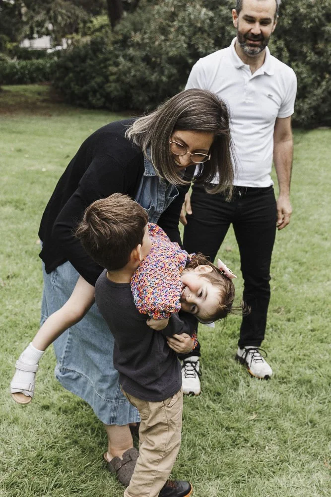 A woman playfully lifting a young girl onto her back while a boy holds her, and a man watches smiling in an outdoor park.