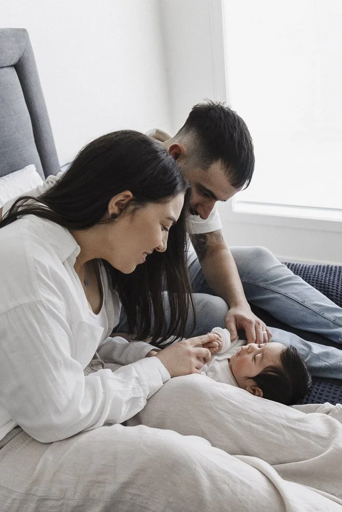 A family of three, including a woman, a man, and a young girl, sitting on a bed in a bright room, engaging happily with the girl.