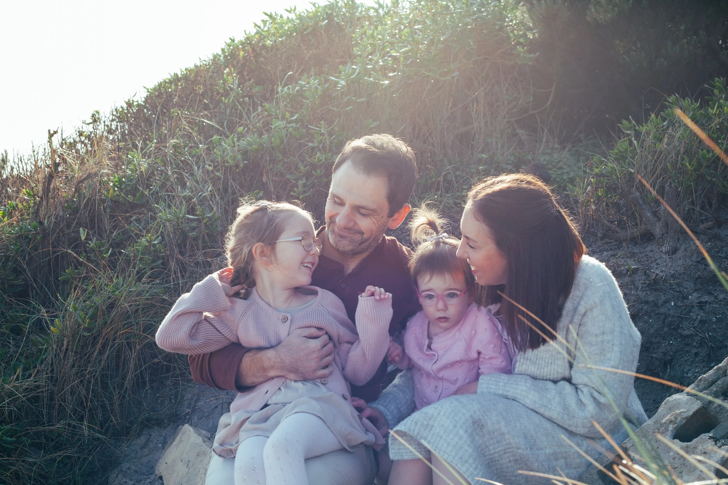 A family of five sitting outdoors on the ground, surrounded by grass and shrubs, enjoying a sunny day and sharing a happy moment together.