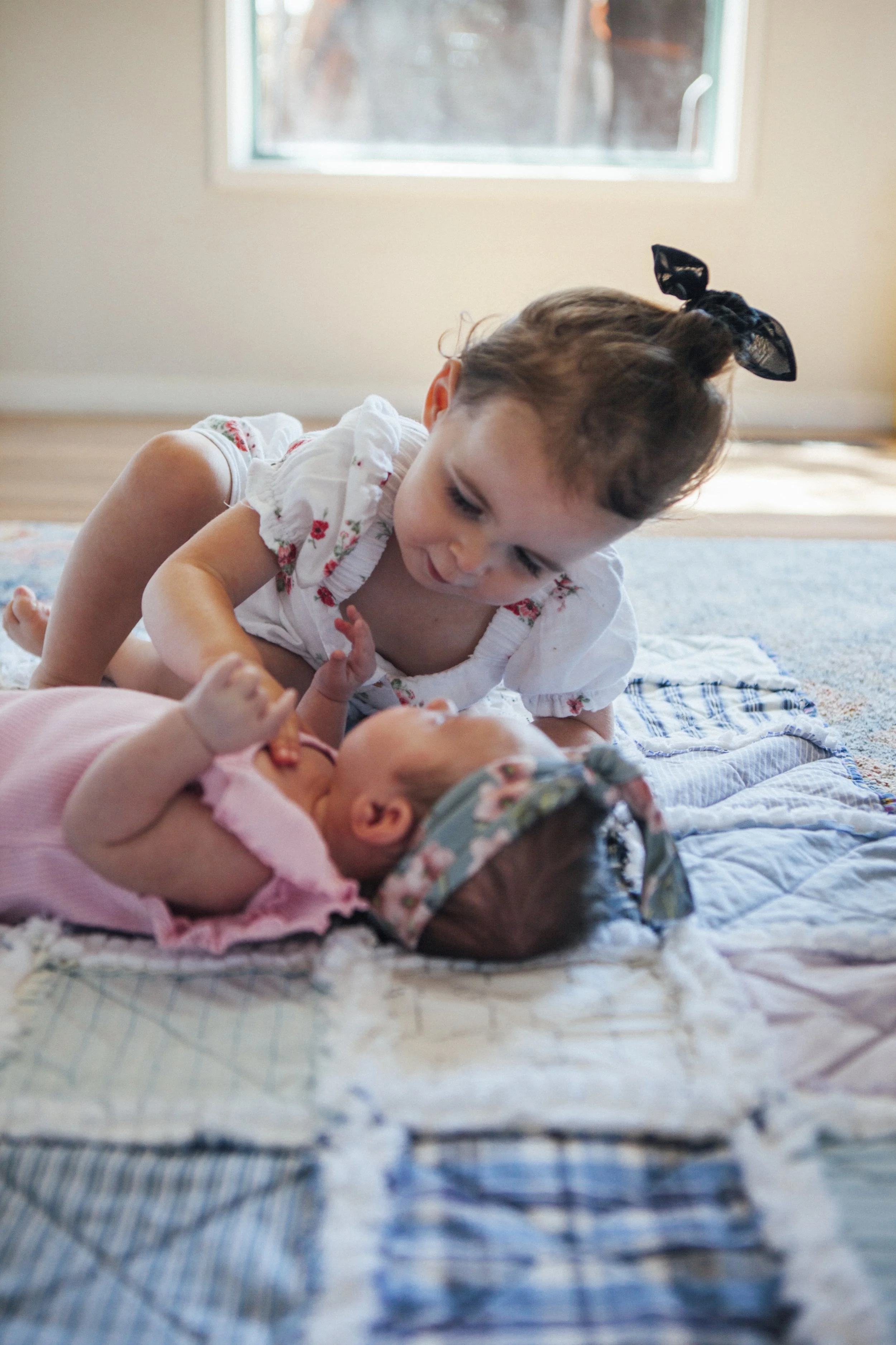 A young girl with a black bow in her hair plays with a baby lying on a quilted blanket on the floor.