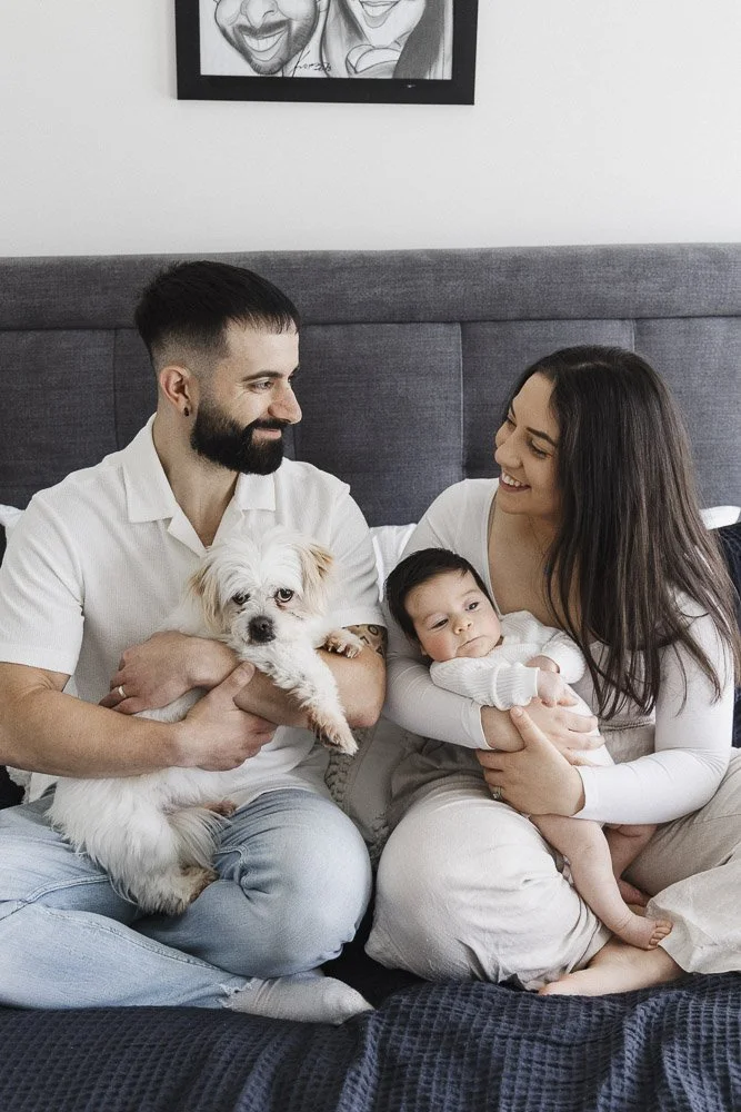 A family of three sitting on a bed with their dog and baby, smiling and looking at each other in a cozy bedroom.