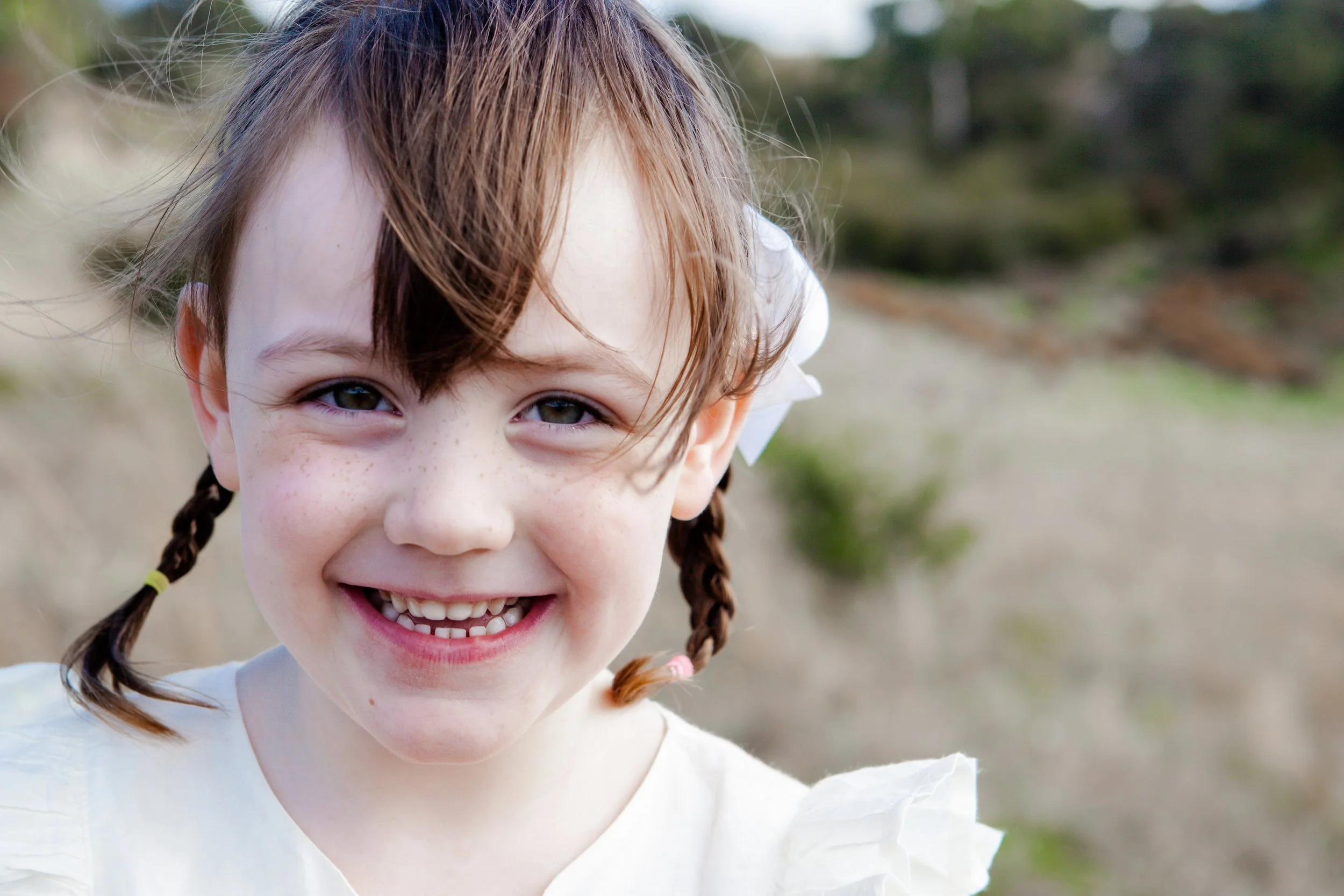 Close-up of a smiling young girl with brown hair in braids, freckles, and a white bow, outdoors in a natural setting.