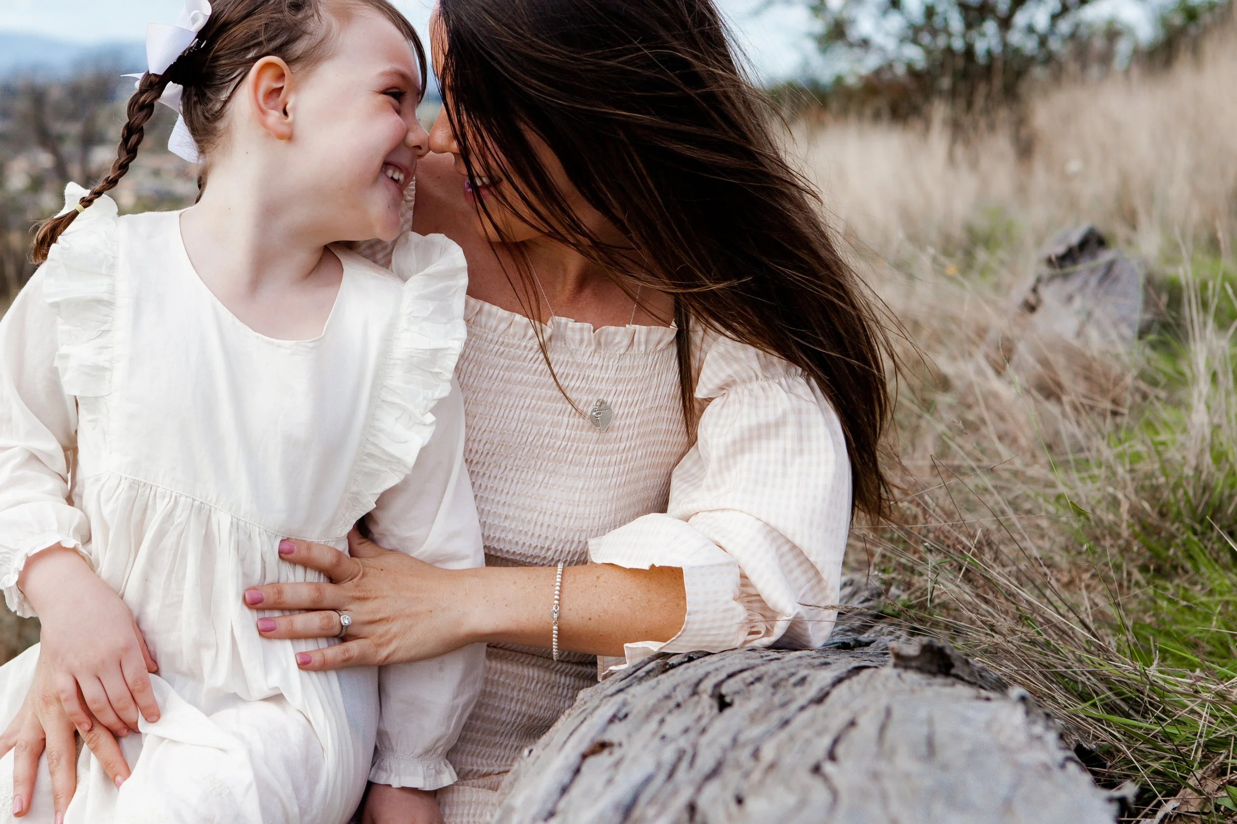 A woman and young girl are close to each other, smiling, with their foreheads touching outdoors in a grassy area with a log in the foreground.