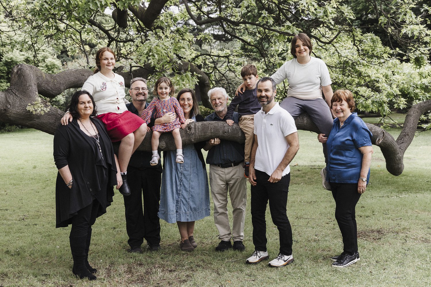 A multi-generational family poses outdoors around a large tree with green leaves. Some family members are sitting on or leaning against the tree branch, while others stand in front. Everyone is smiling and enjoying the gathering.