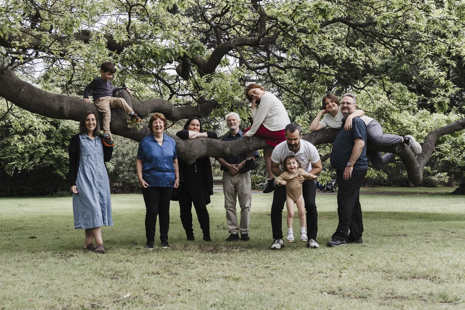 A group of people, including children and adults, are gathered outdoors around a large tree. Some are sitting on or hanging from the tree's branches, while others are standing on the ground. The scene is in a park with green grass and trees in the ba
