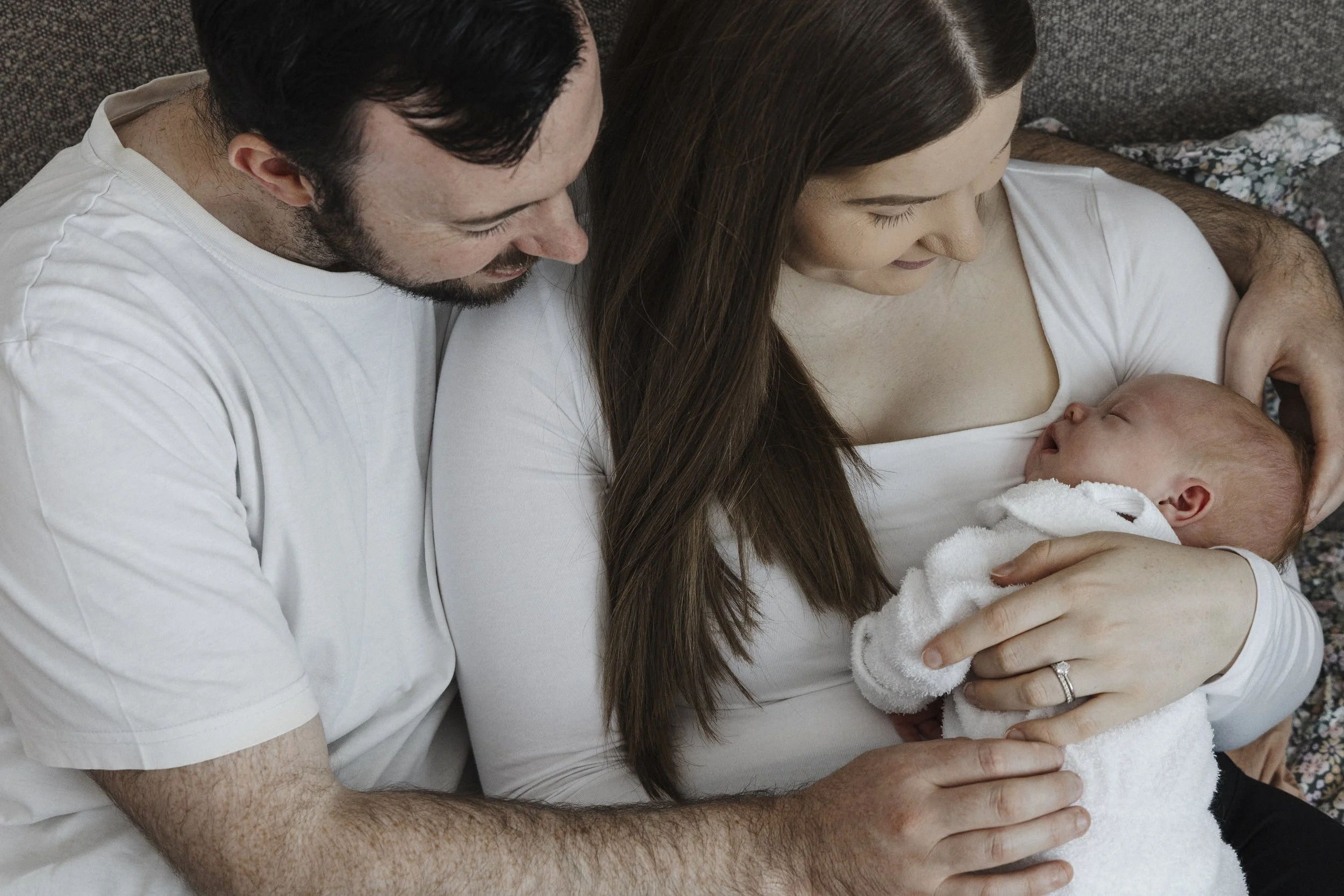 A man and woman sitting on a couch with a baby. The woman is holding the baby and looking at it, while the man is leaning in and looking at the baby. The woman is wearing a white top and the man is wearing a white shirt. The baby appears to be sleepi