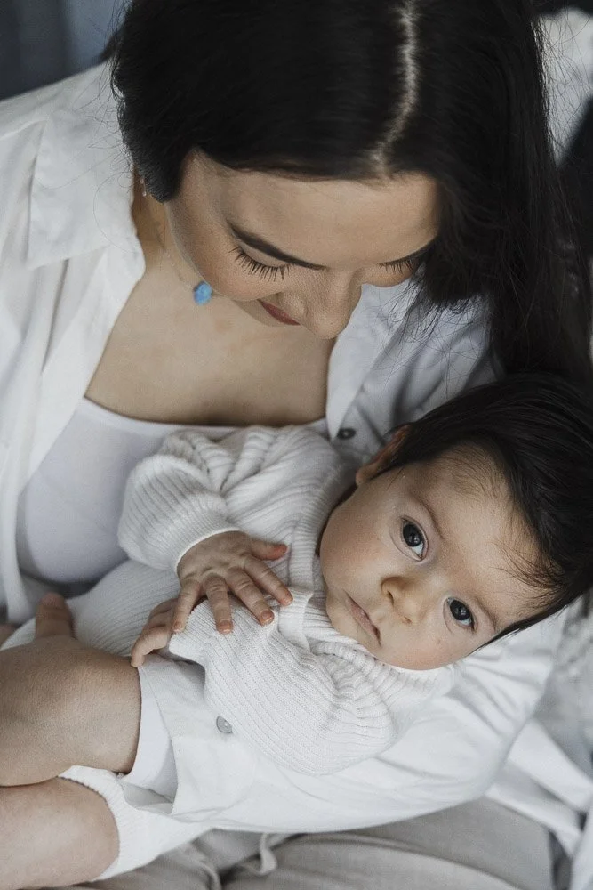 A woman gently holding and looking at a baby with dark hair and big eyes, lying in her arms, indoors.