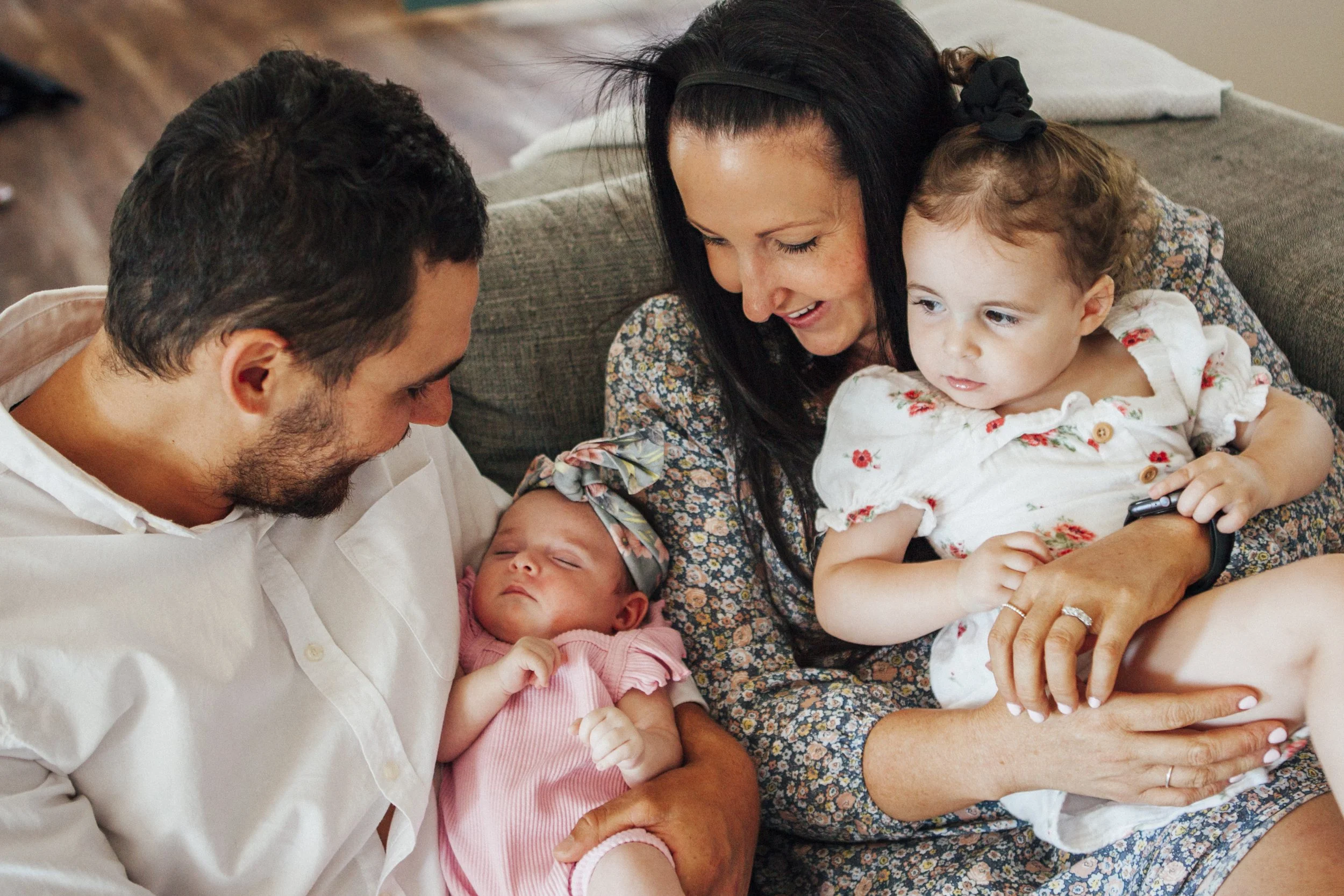 A family of four sitting on a couch: a man, a woman, a young girl, and a baby. The man is holding the sleeping baby, and the woman, wearing a floral dress, is holding the young girl who is looking at a phone, while the man looks at the baby.