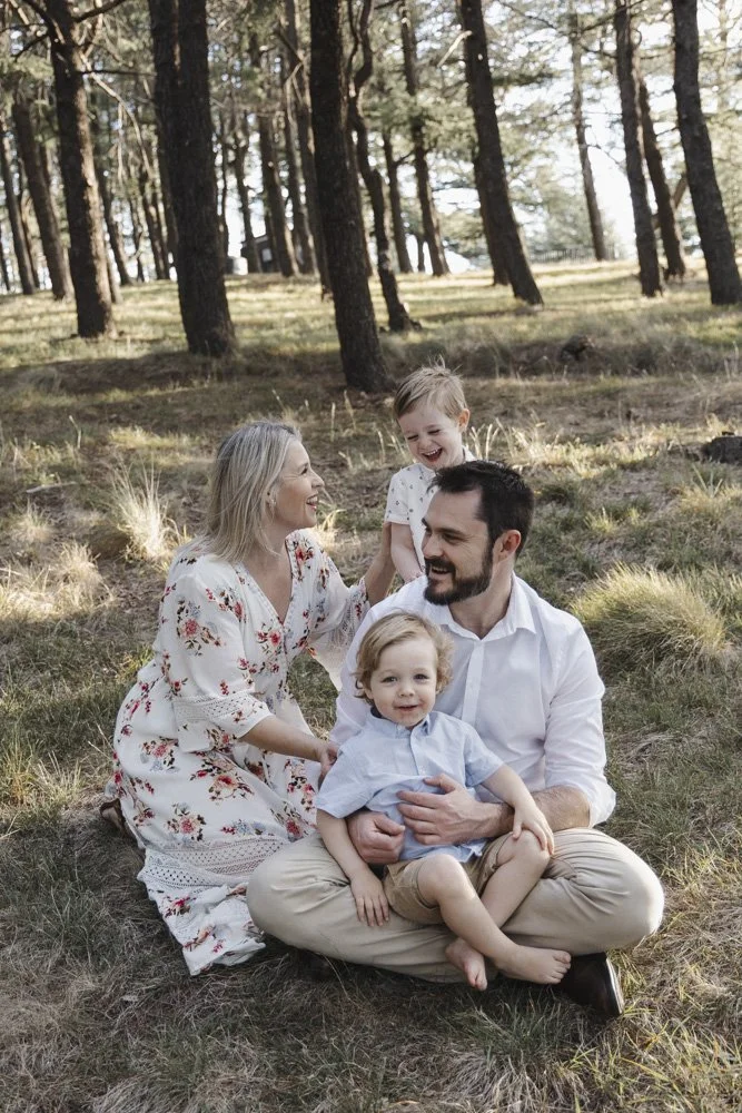 A family with two children sitting on the grass in a forest, smiling and enjoying each other's company.