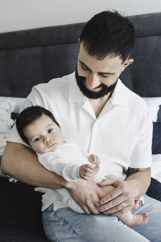 A father with a beard sitting on a bed, holding an infant child in his arms and looking down at the child with a gentle smile.