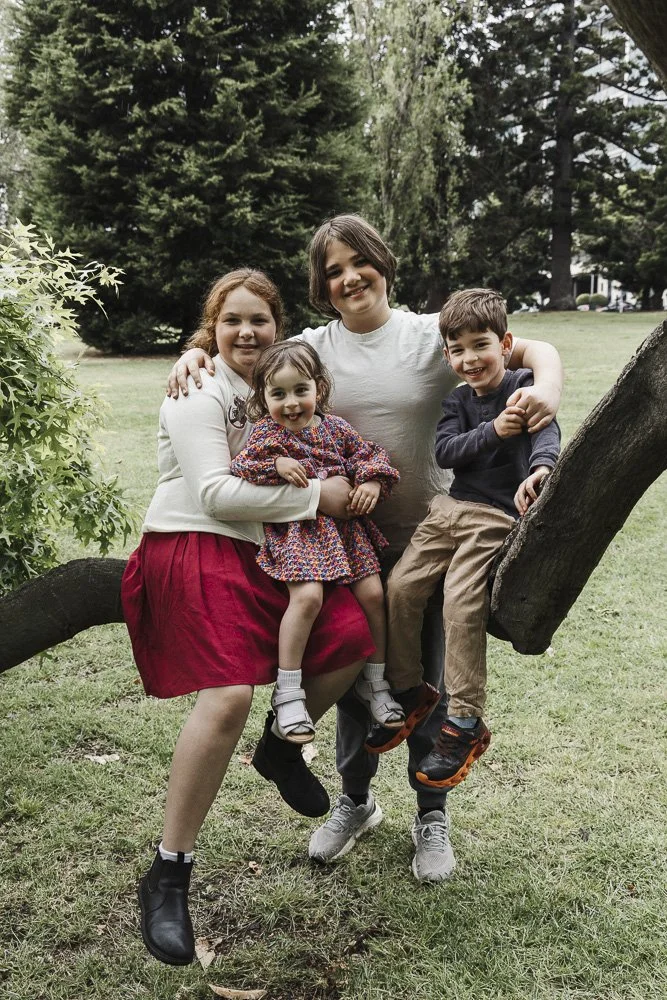 A family of four, including children, sitting on a tree branch in a park with green grass and tall trees in the background, smiling at the camera.