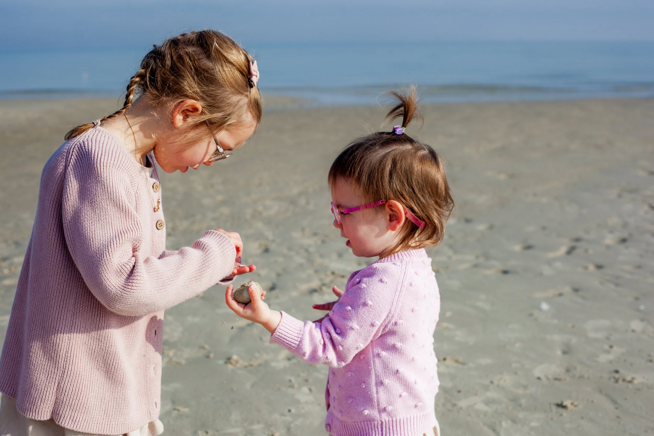 Two young girls on a sandy beach, exchanging a small object, with the ocean in the background.