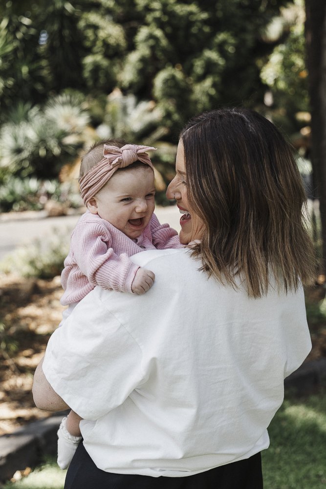 Mother holding smiling baby girl in a park with trees and greenery in the background.