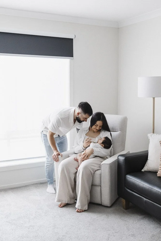 A woman is sitting in a beige armchair, holding a breastfeeding baby, while a man stands beside her, looking at the baby in her arms. They are in a bright, minimalist living room with white walls, a window with a shade, and modern furniture.