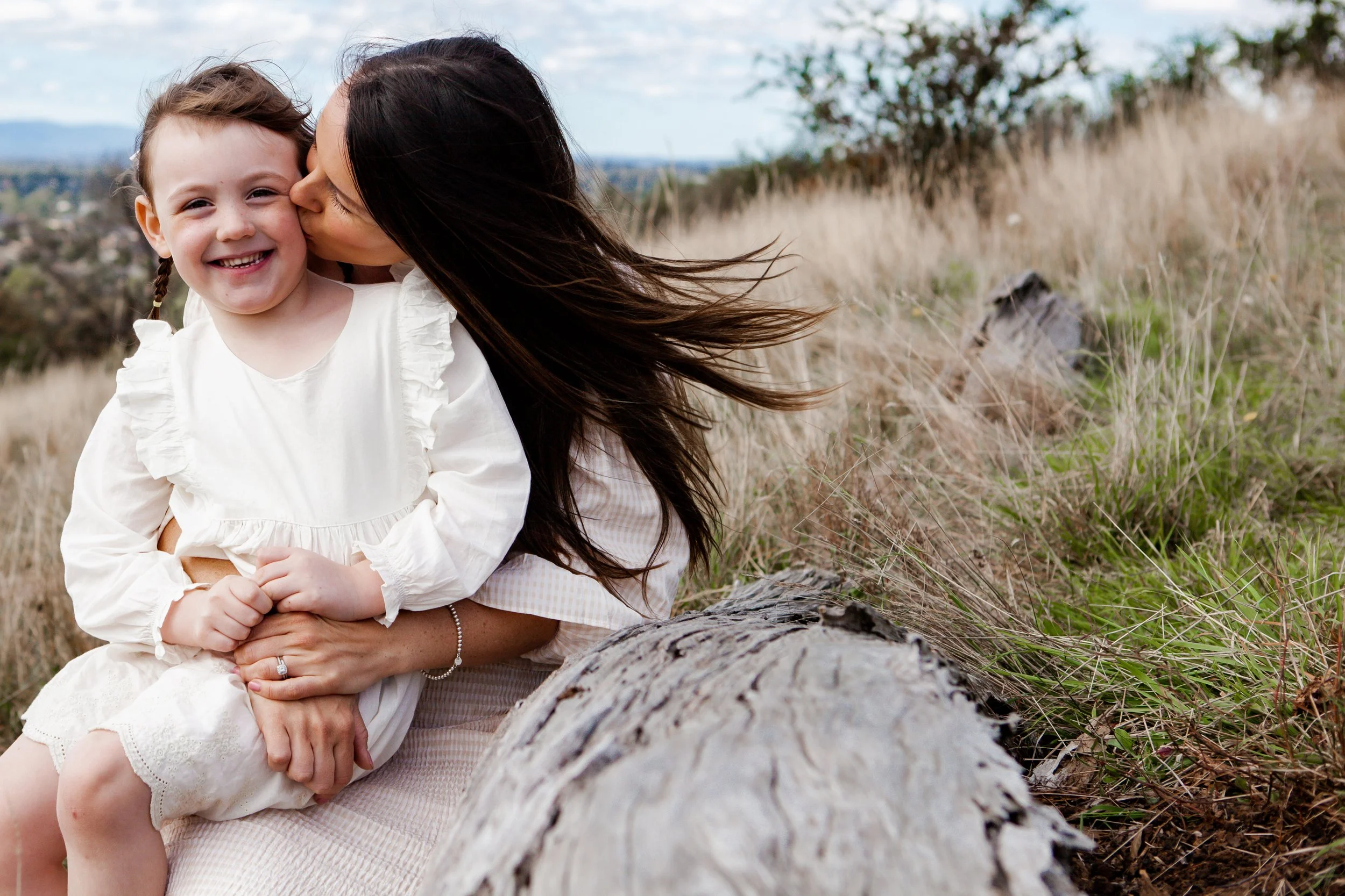 A woman is kissing a smiling young girl on the cheek outdoors in a grassy field with a fallen log and trees in the background.