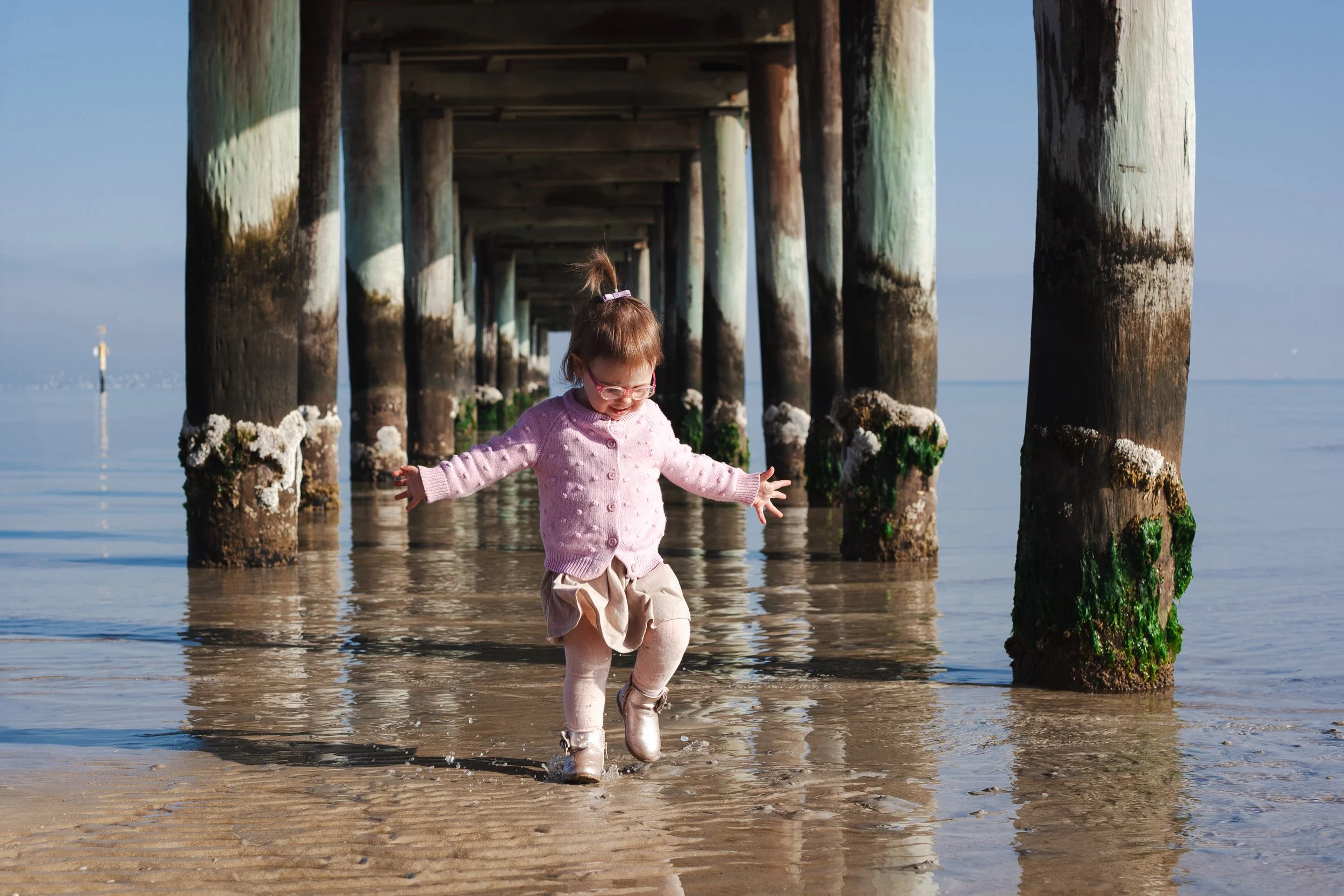 A young girl in pink sweater and beige skirt playing in shallow water under a pier, with large wooden pilings covered in green algae.