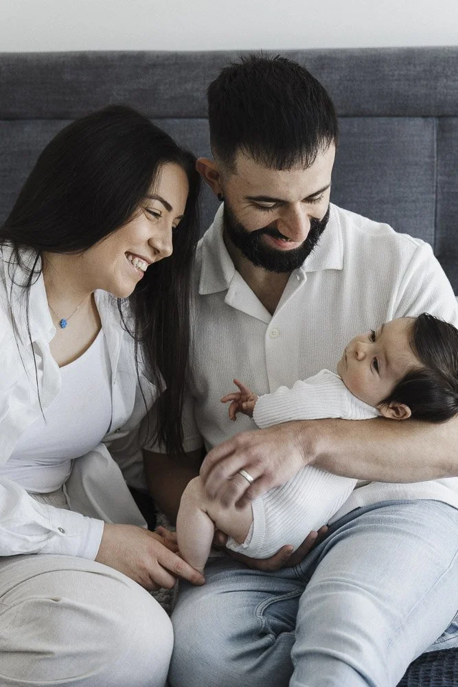 A happy family, a woman, a man, and a baby, sitting together on a bed, smiling and looking affectionately at each other.