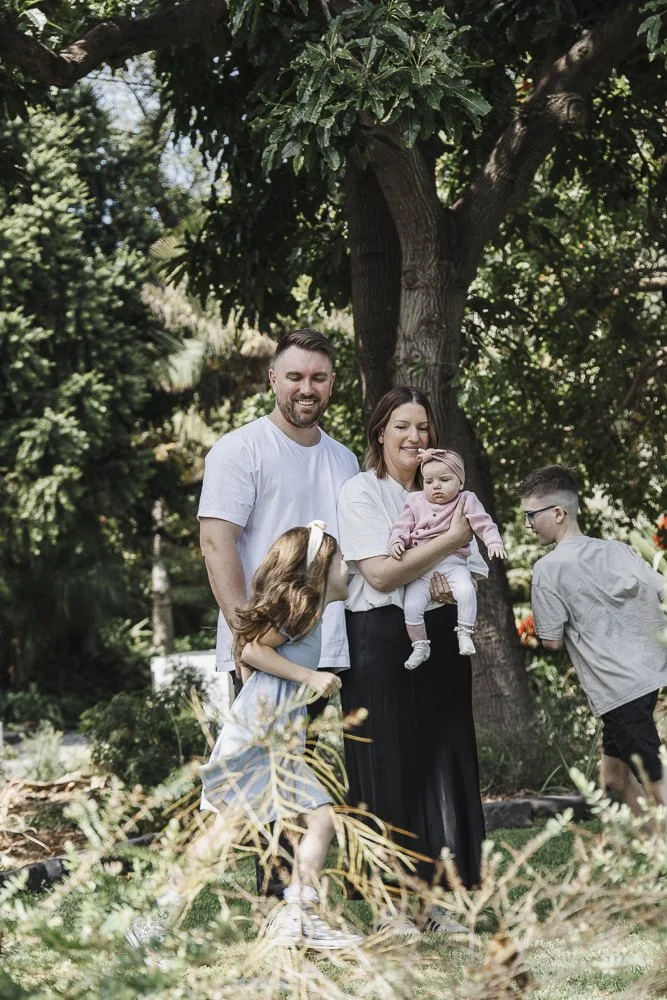 A family of four outdoors under a large tree, with a man, woman, and two children, smiling and enjoying a sunny day in a lush garden.