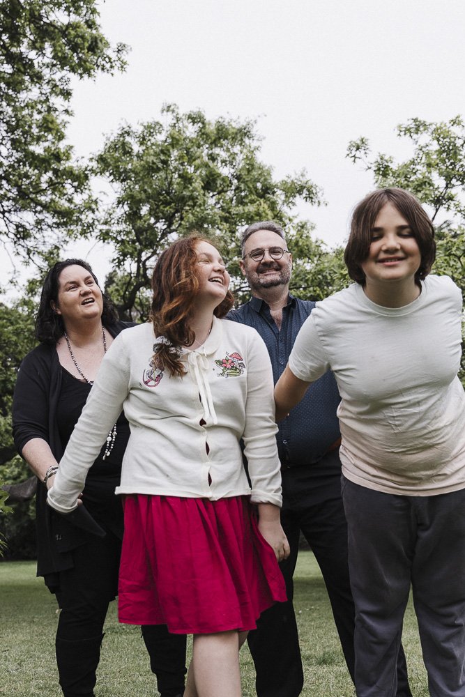 A group of four people, two women, one man, and a young girl, standing outdoors on grass with trees in the background, smiling and enjoying the moment.