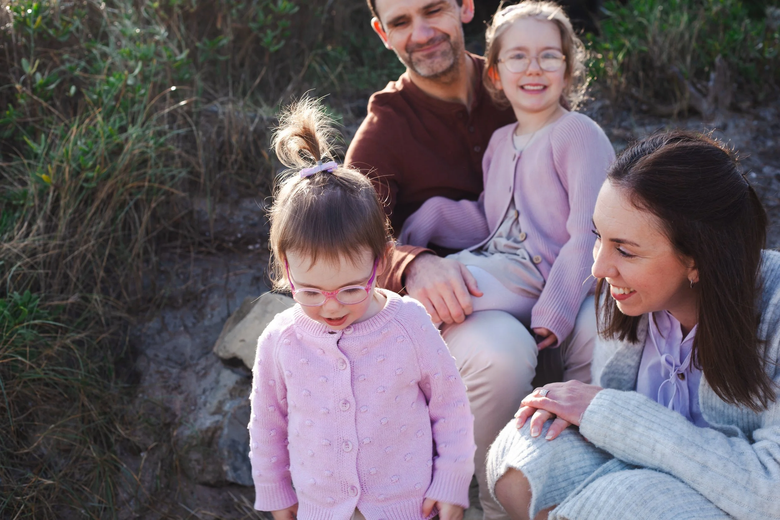 Family of four, including two young girls and two adults, sitting outdoors near grass and rocks, smiling and enjoying the moment.