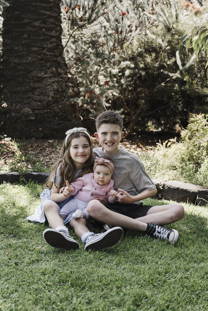 Three children sitting on grass in a park, surrounded by trees and bushes. The older boy and girl, both smiling, are seated with their younger sister, who has a neutral expression, all wearing casual clothing.