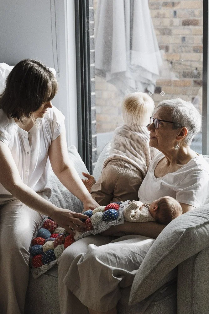 A woman and two children sitting on a sofa with an elderly woman, possibly a grandmother, holding a newborn baby in her arms, near a large window with a brick wall outside.