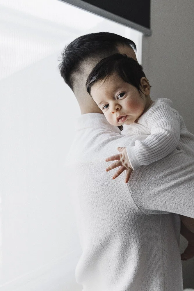A young child with dark hair being held in an adult's arms, looking over the adult's shoulder towards the camera, wearing a white sweater.