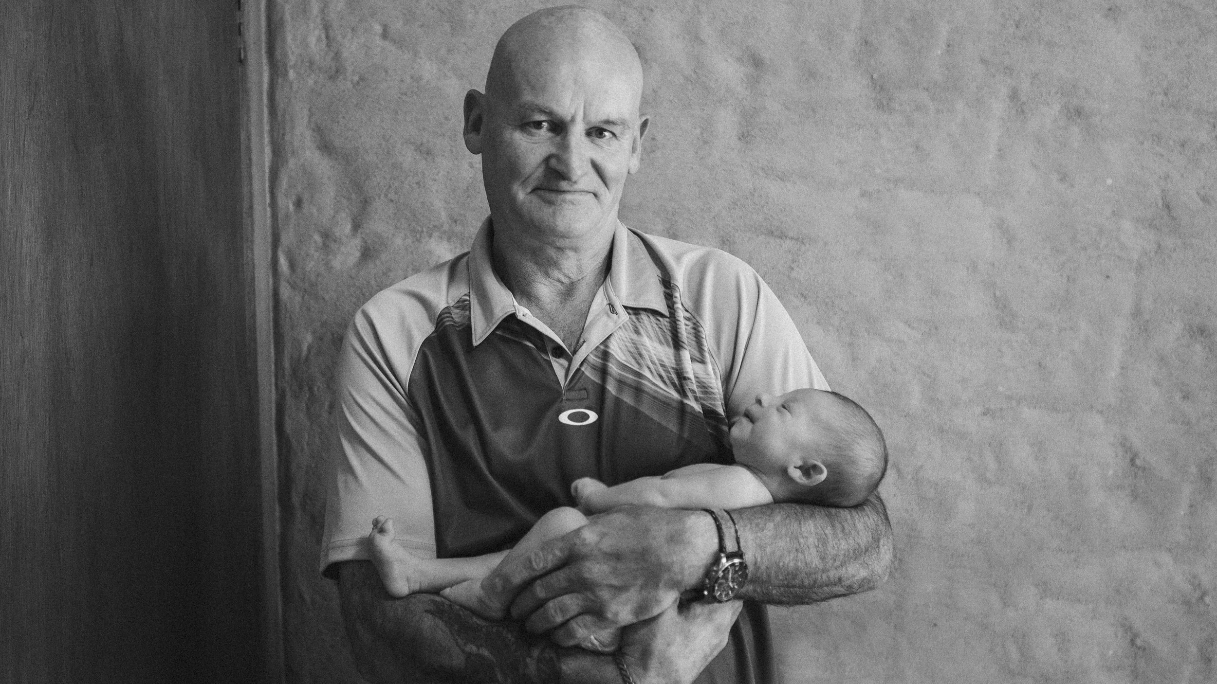 An older man with a serious expression holding a sleeping baby in his arms, standing against a plain textured wall.