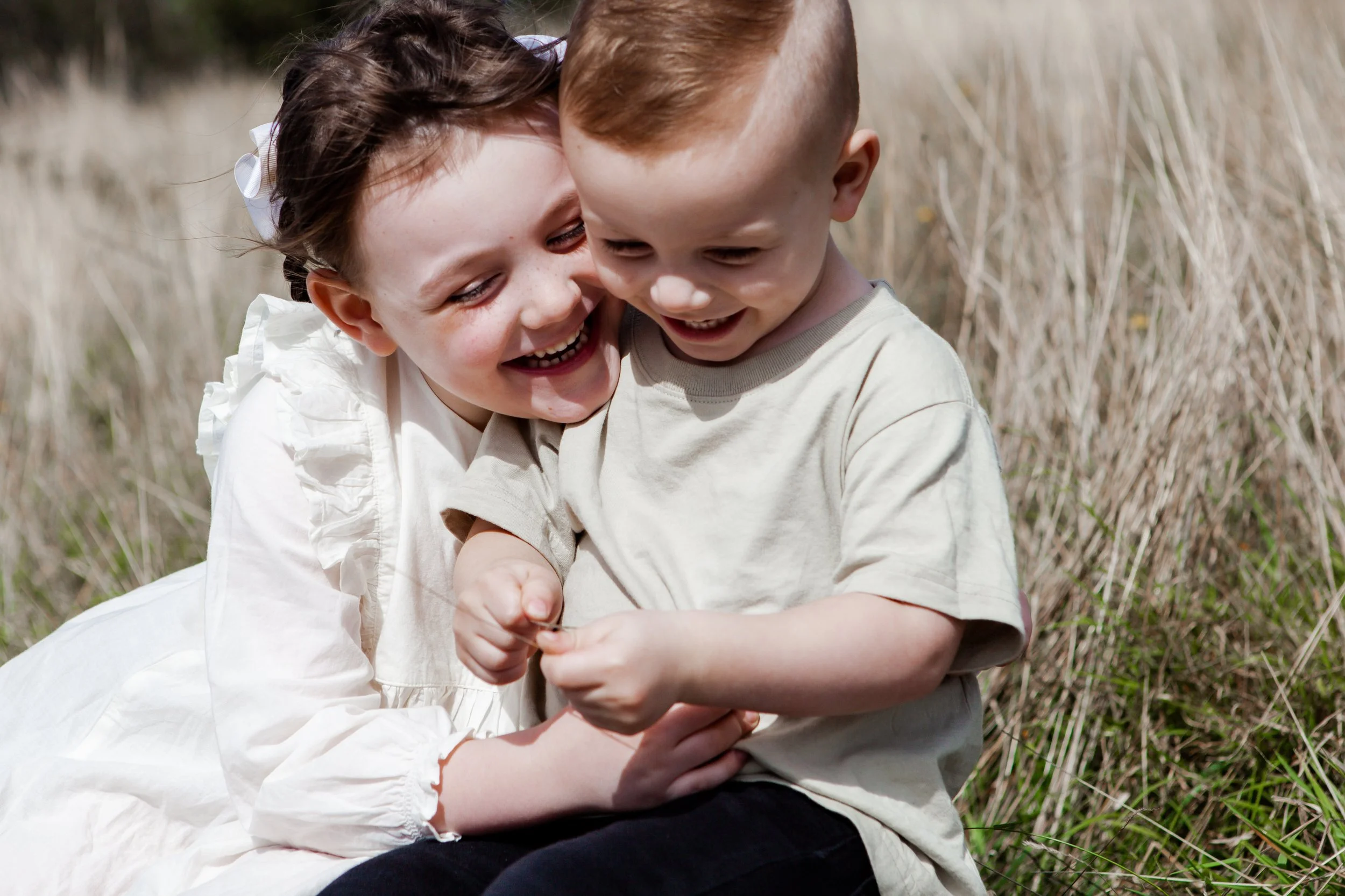 Young girl and boy laughing and hugging in a grassy field.