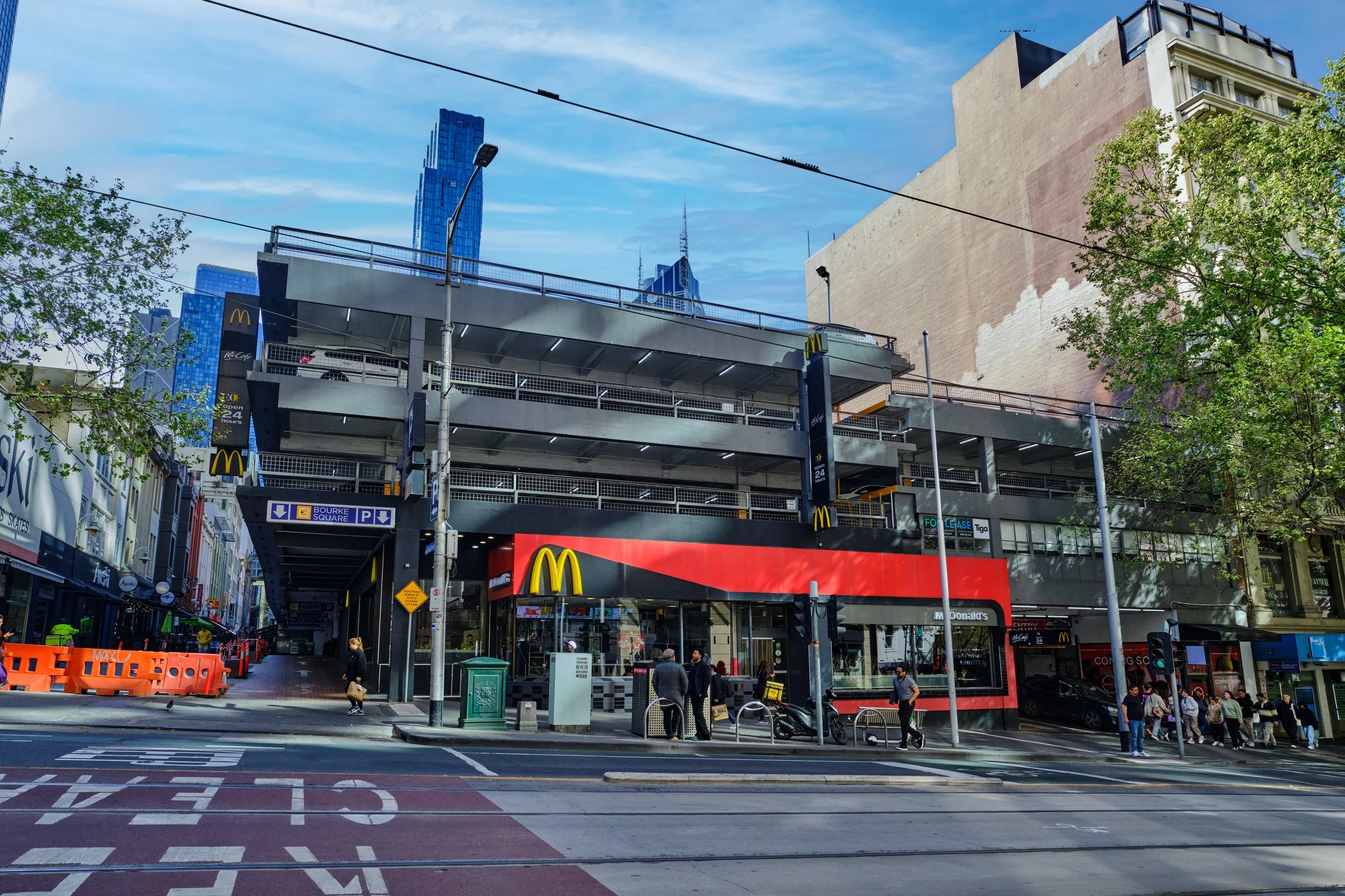 A multi-story parking garage and McDonald's restaurant in an urban area with tall skyscrapers in the background, pedestrians walking on the sidewalk, tram tracks in the street, trees, and street signs.