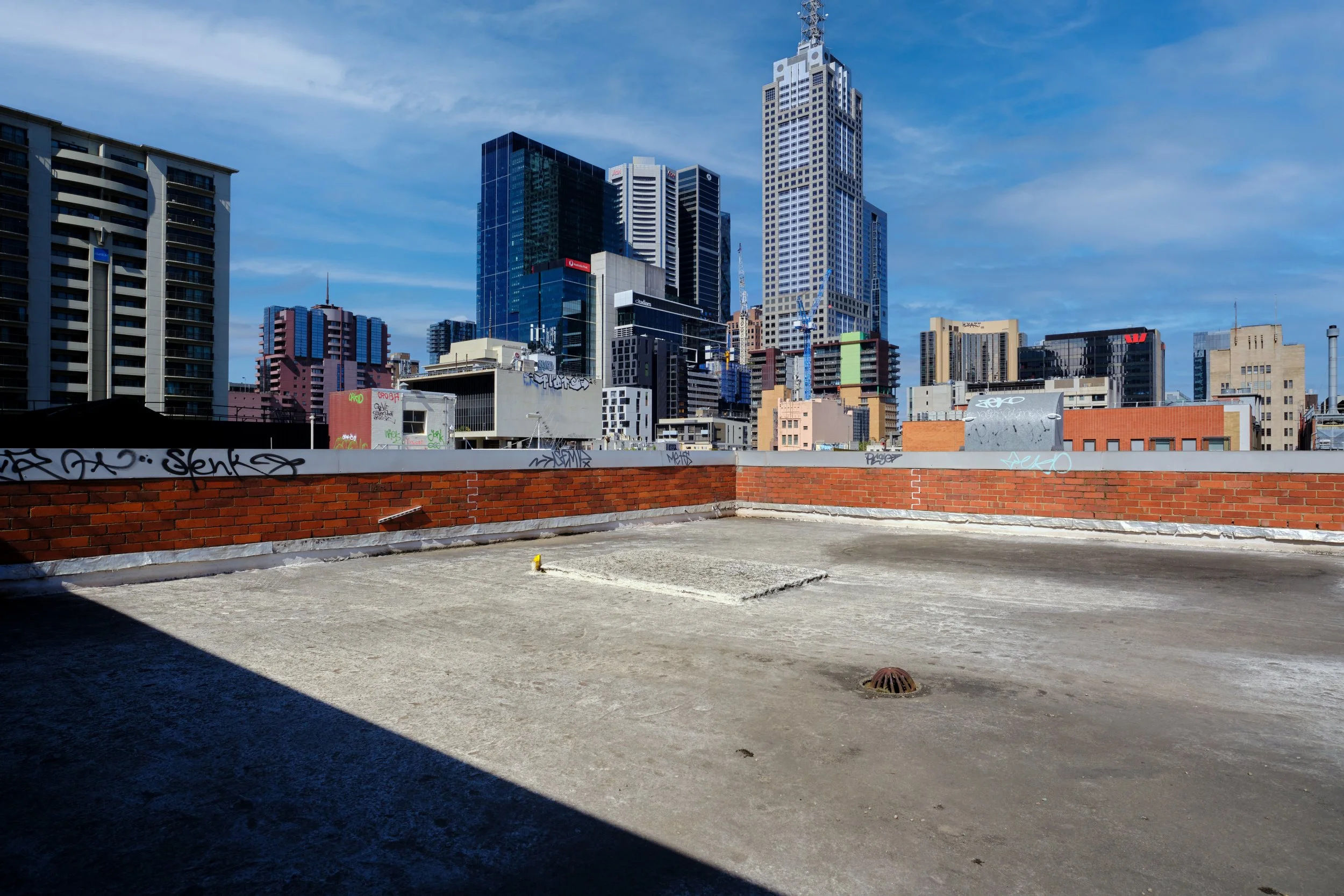 View of city skyline with tall buildings under blue sky, seen from a rooftop with concrete surface, red brick wall, and graffiti.