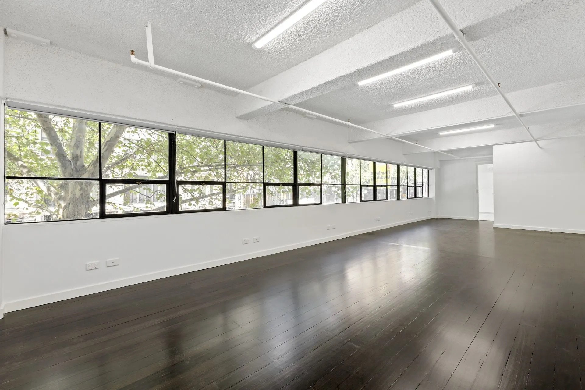 Empty room with large windows, white walls, and dark wooden floors, likely an office or studio space.