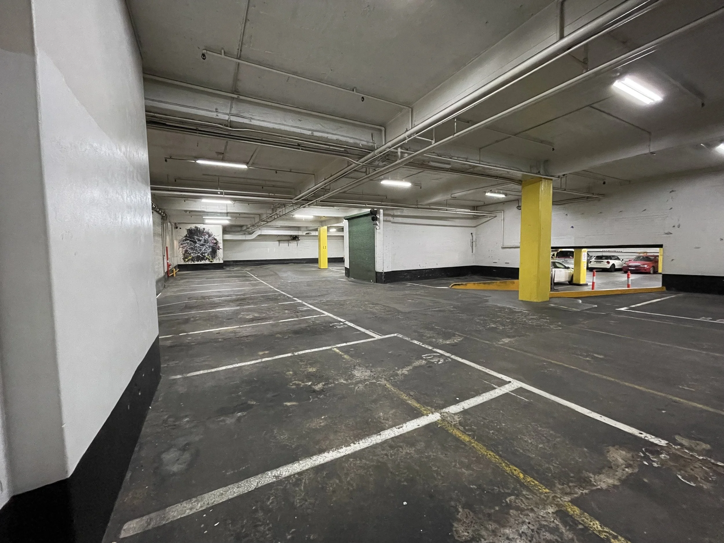 Empty underground parking garage with marked parking spaces, concrete walls, ceiling pipes, and a few parked cars visible in the background.