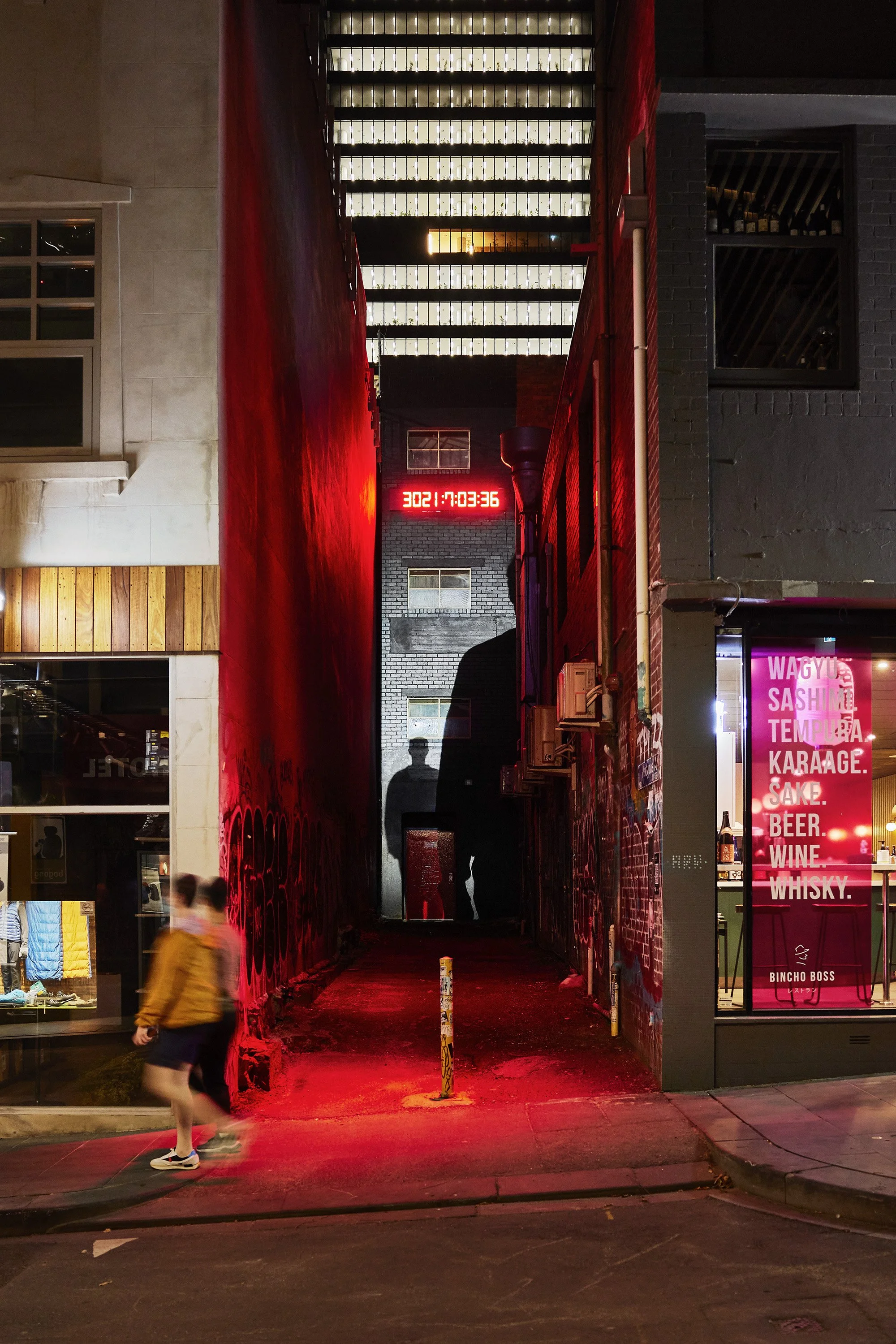 A nighttime urban scene showing a narrow alleyway between two buildings with vibrant red lighting. A digital clock displaying '3021:7:03:36' is mounted on the back wall, and a silhouette of a person is reflected on a glass surface in the alley. To the right, a storefront with neon signage lists foods and beverages like Wagyu, sashimi, tempura, karaage, cake, beer, wine, and whisky.