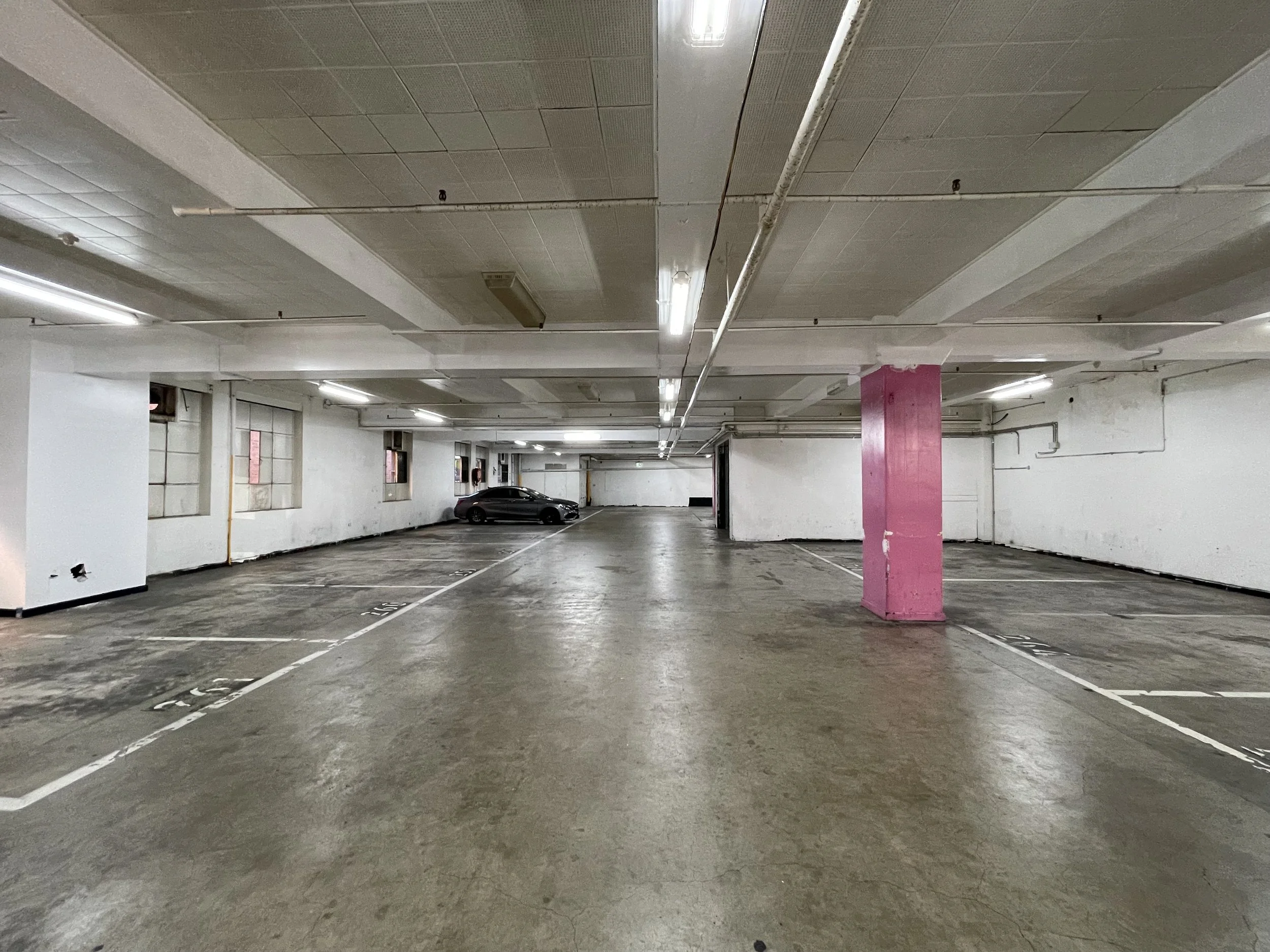 Empty underground parking garage with a single black car parked in the background, white walls, pink column, fluorescent lighting, and visible pipes.