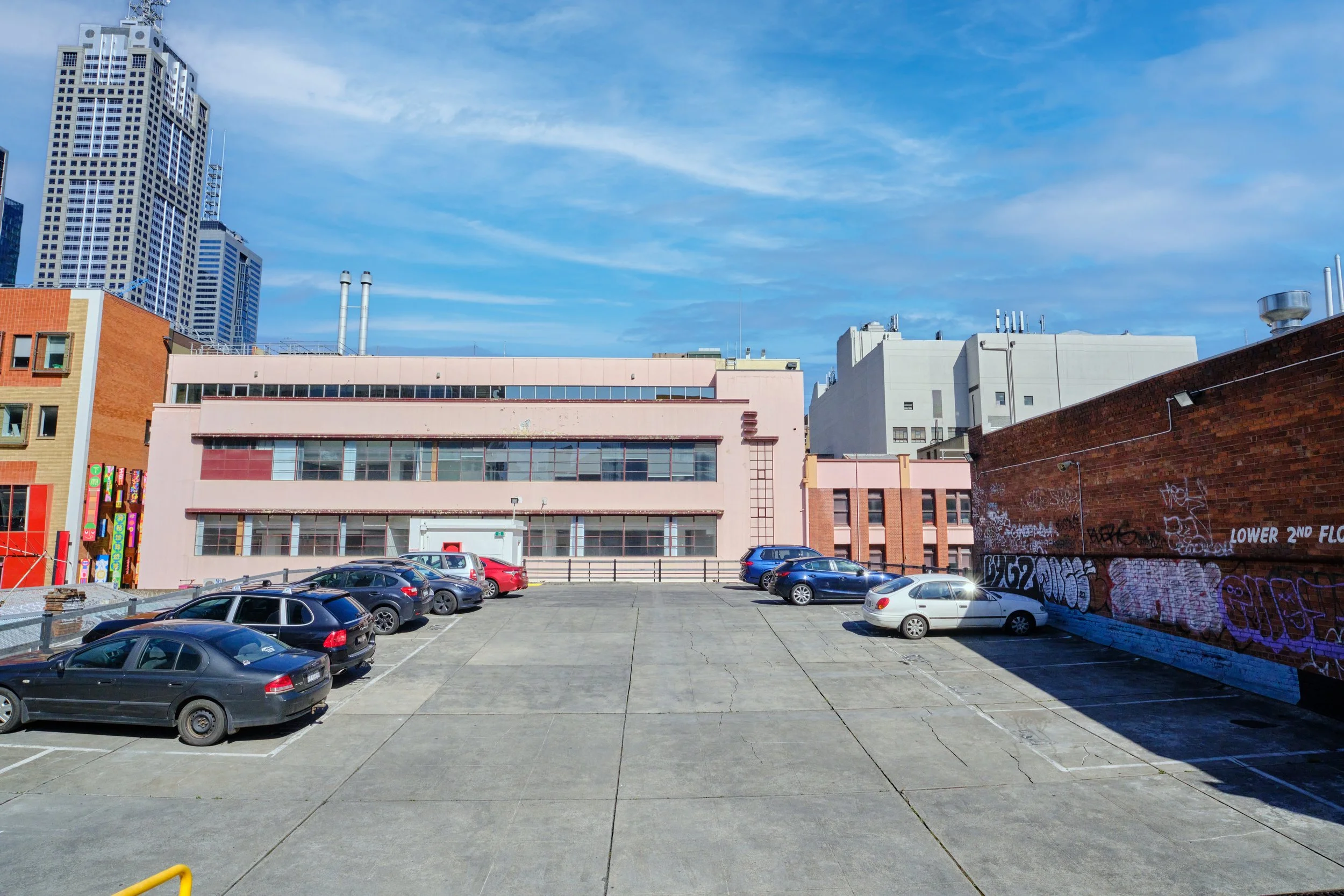 Empty city parking lot with parked cars, colorful buildings, and tall skyscrapers under a blue sky with white clouds.