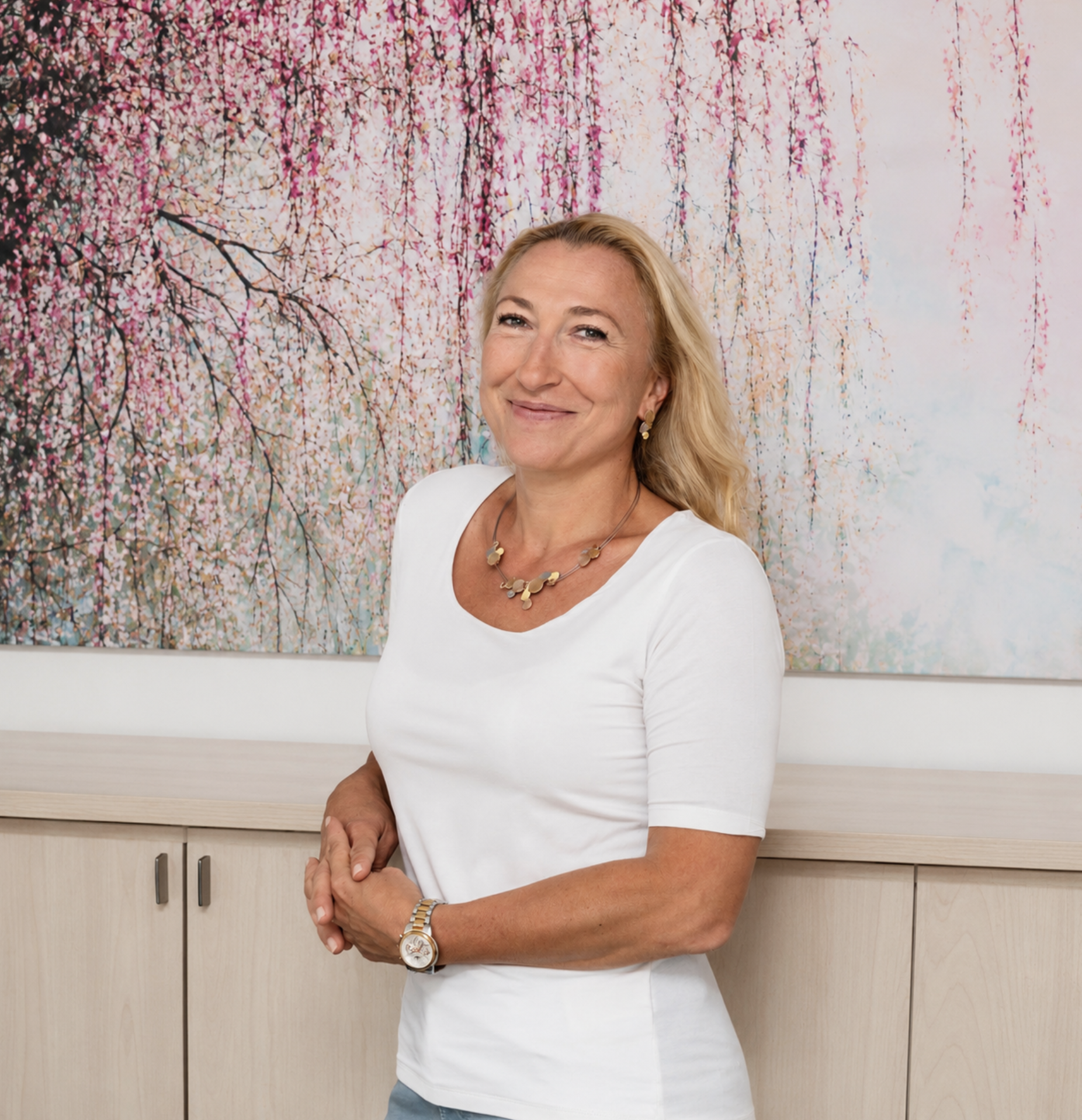 Eine lachende Frau mit blonden, welligen Haaren trägt eine weiße T-Shirt und eine goldene Uhr und eine Halskette, steht vor einem Himmelblauen und rosa Blüten hintergrund.
