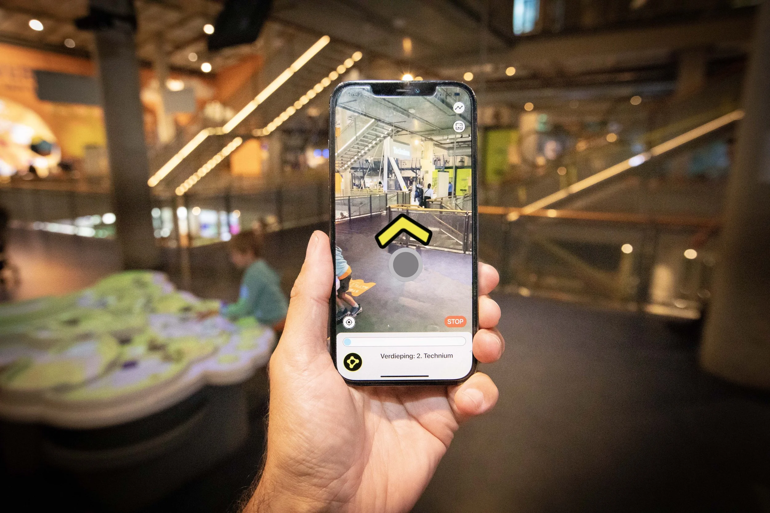 Hand houdt een smartphone vast met de eZwayZ-app open. Op het scherm is een binnenruimte te zien met een looproute en een gele richtingspijl. Onderaan staat de tekst “Verdieping 2. Technium”. Op de achtergrond is dezelfde ruimte onscherp zichtbaar.