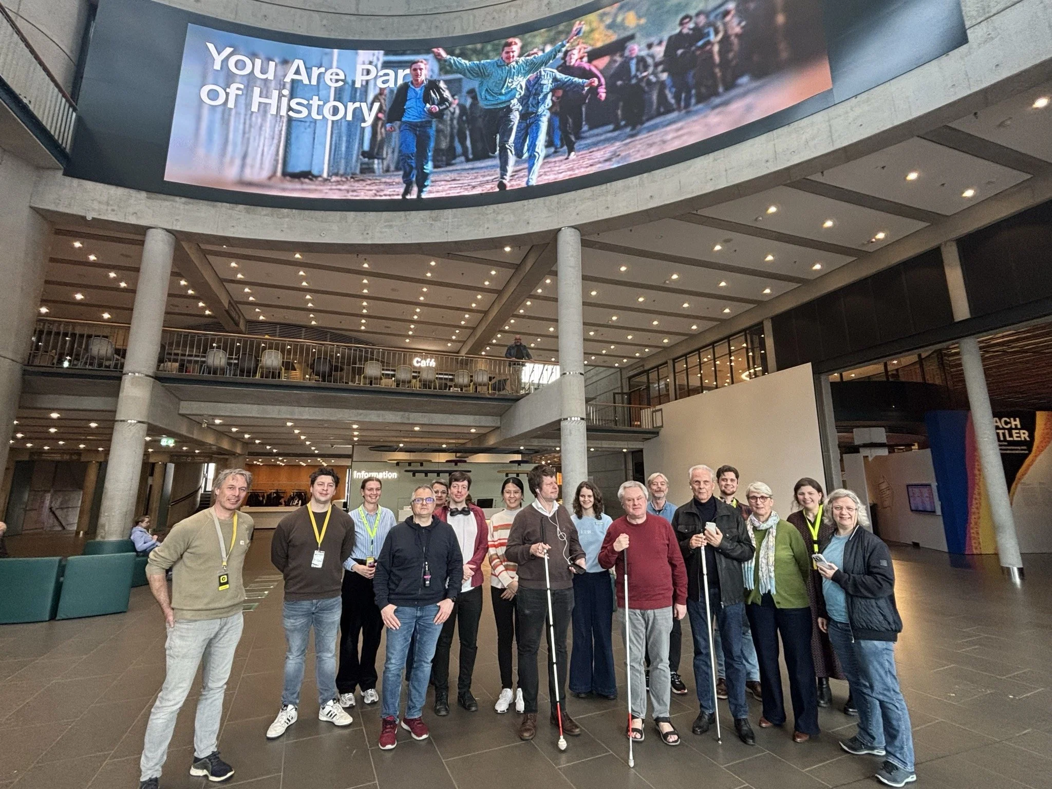 Grote groep mensen poseert in een museumhal, van wie enkelen met een blindenstok, met op een scherm de tekst You Are Part of History.