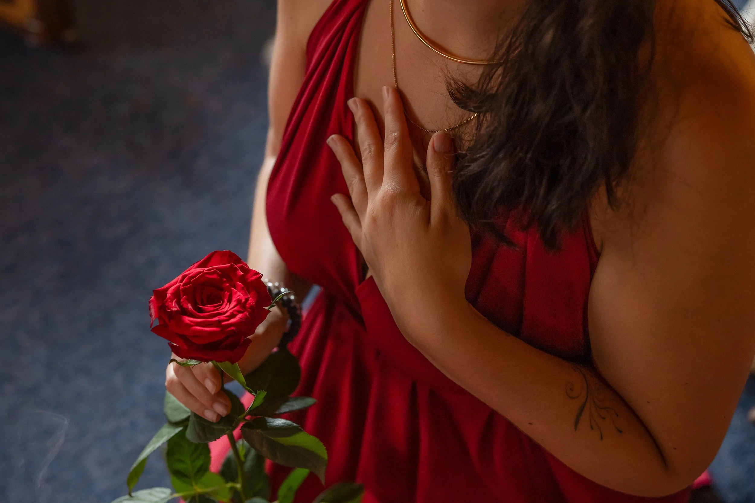A woman in a red dress holding a red rose close to her waist, with her left hand touching her chest, and a tattoo on her left forearm.