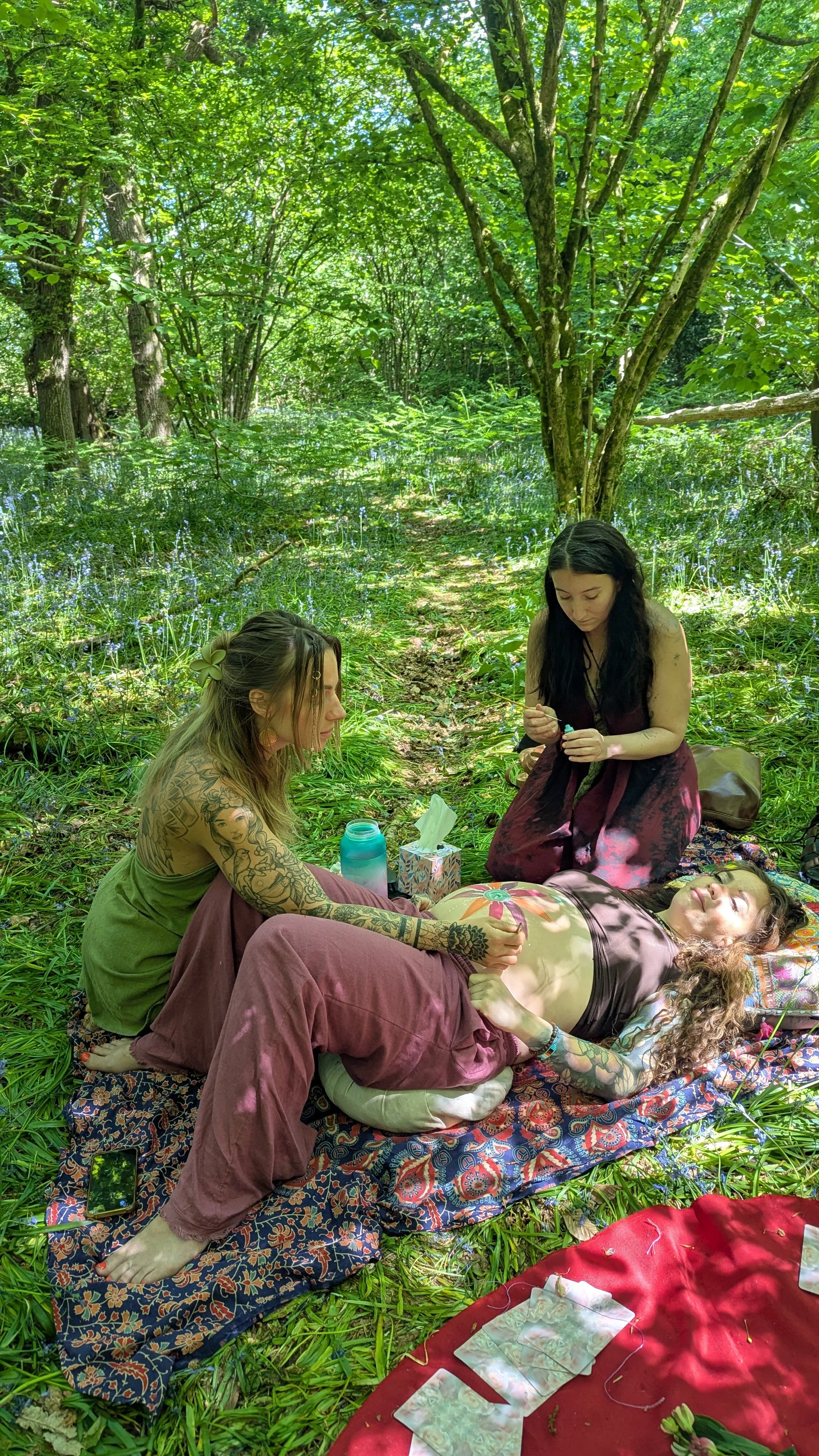 Three women with tattoos participating in a spiritual or healing ritual in a lush green forest, with one woman lying on a blanket and two women sitting nearby, one holding the other's hand.
