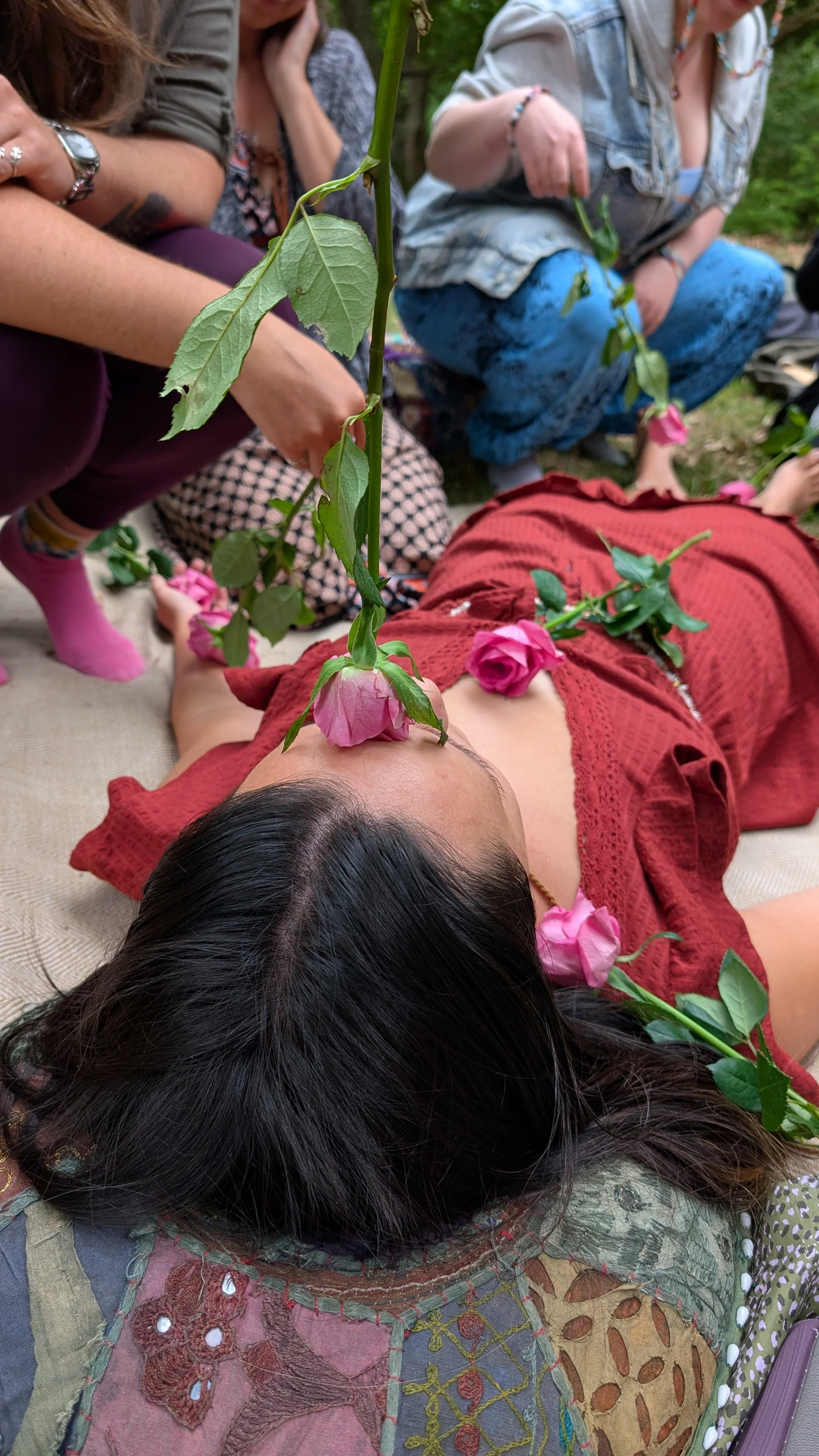 A woman lying down with pink roses on her chest and face, surrounded by people outdoors, some sitting and one with a flower held above her face.