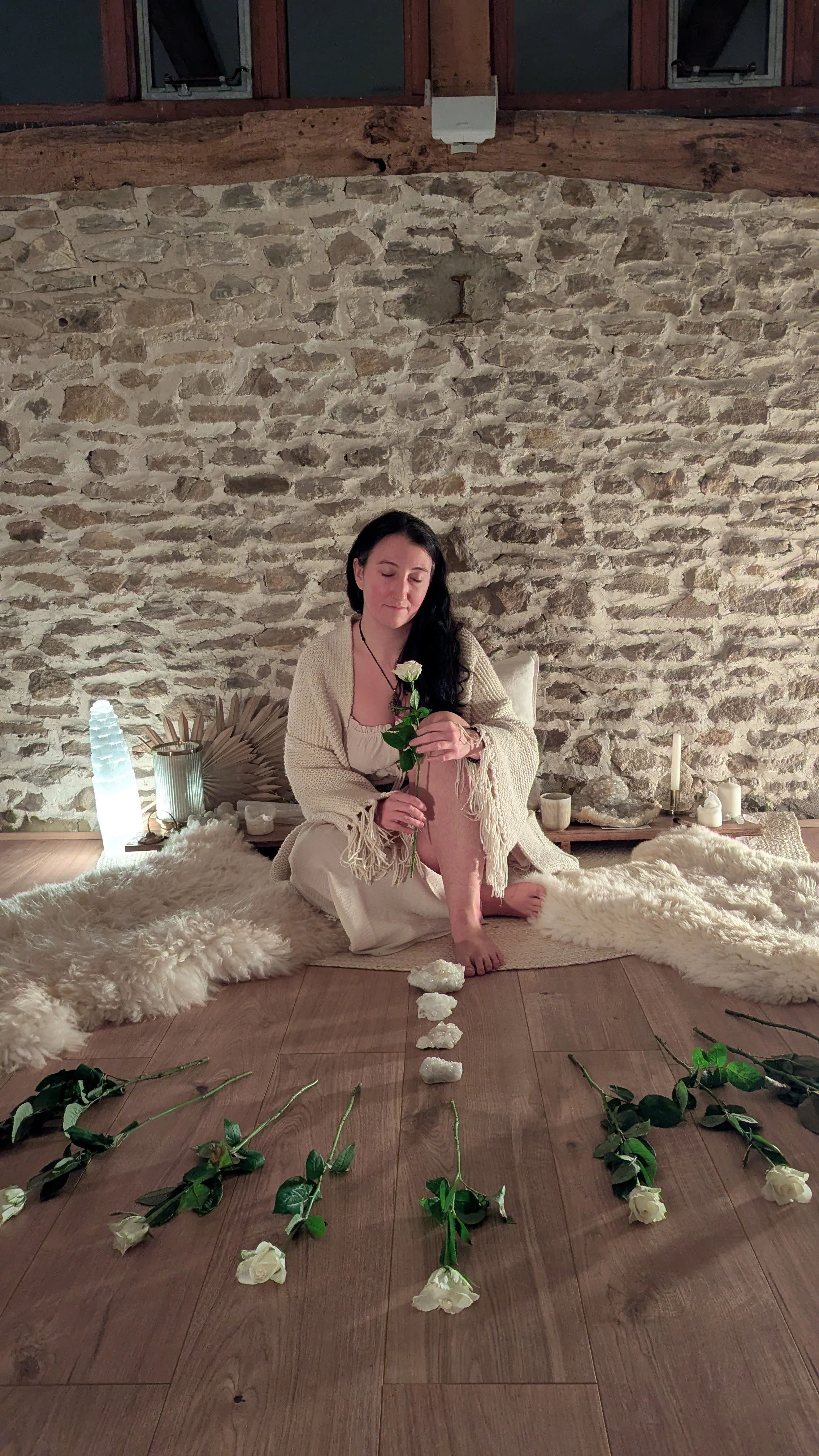 A woman with long dark hair sitting on the floor in front of a stone wall, holding a white rose, surrounded by candles, candles, a fur rug, and flowers arranged in a symmetrical pattern on the wooden floor.