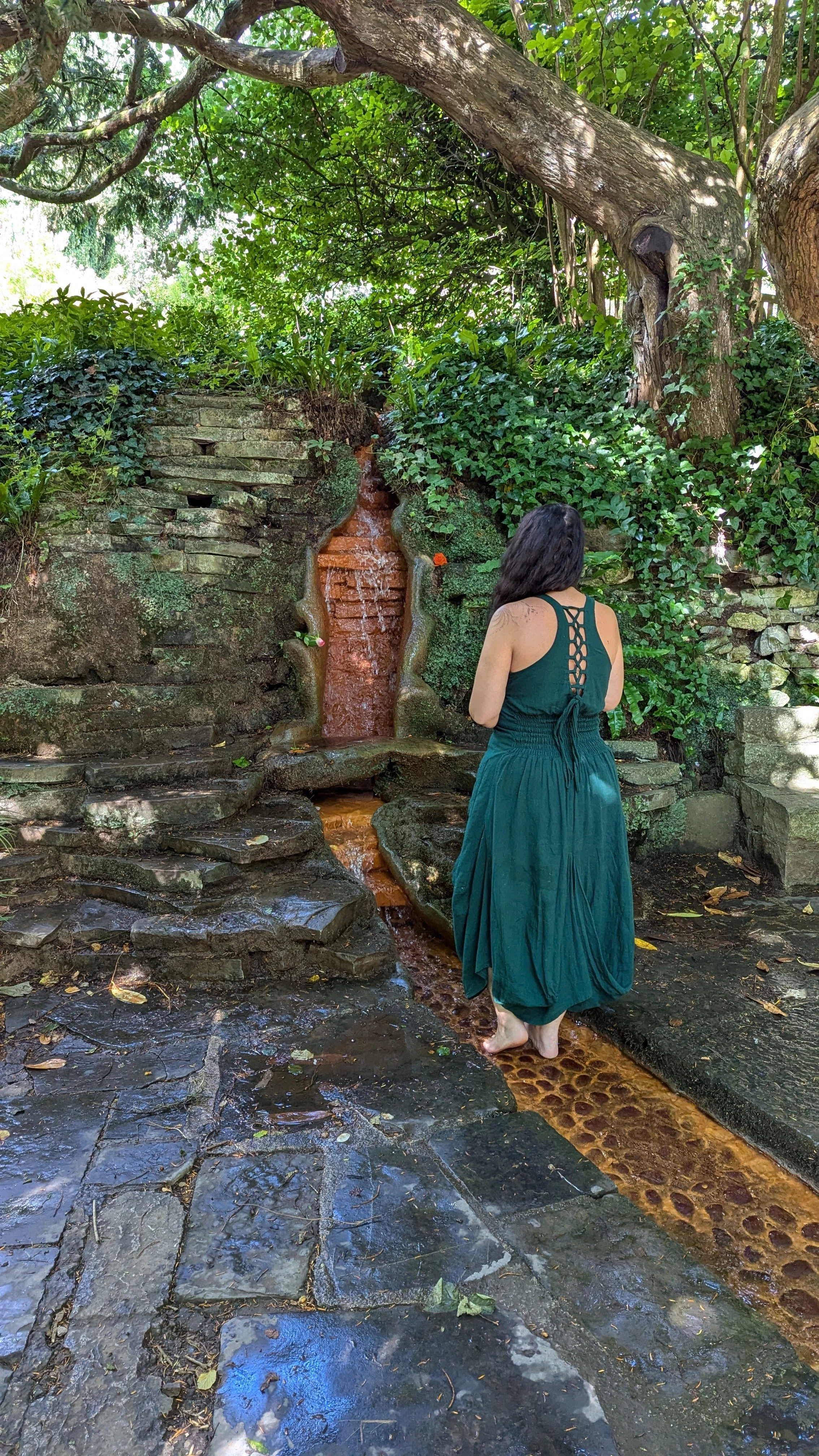 A woman in a teal dress with a lace-up back walks barefoot near a small water fountain in a lush, green garden.