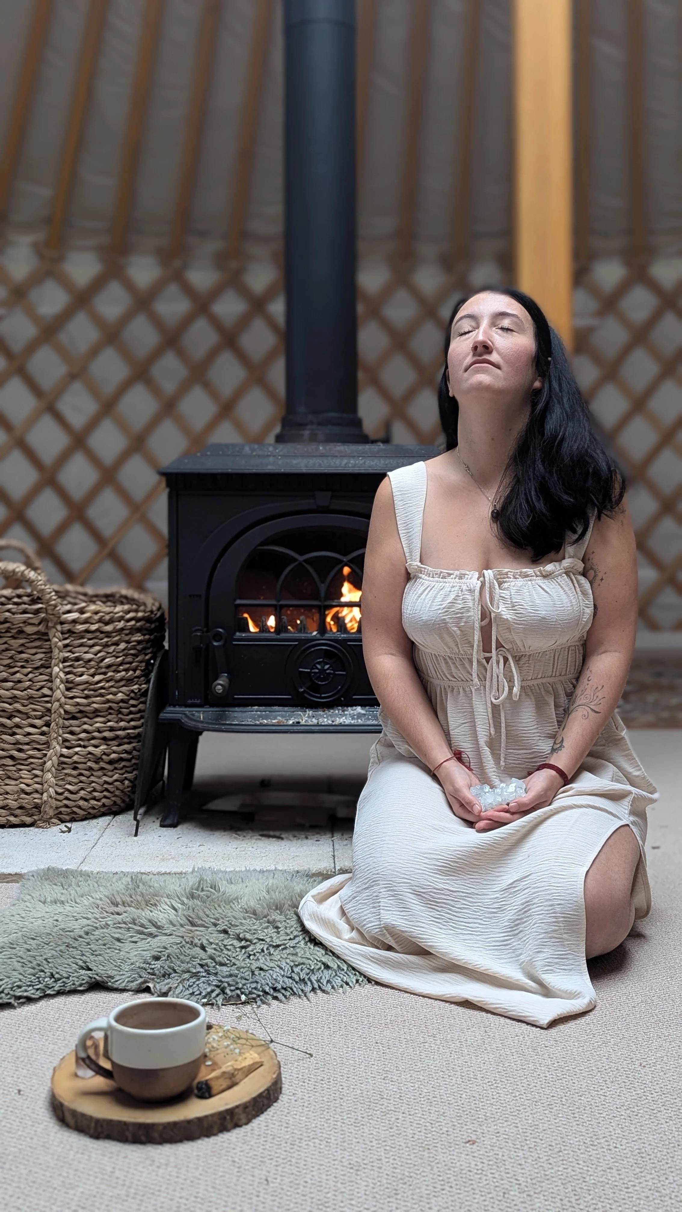 Woman sitting by a fireplace in a cozy yurt, holding crystals, with a cup of coffee on a wooden slice in front of her.