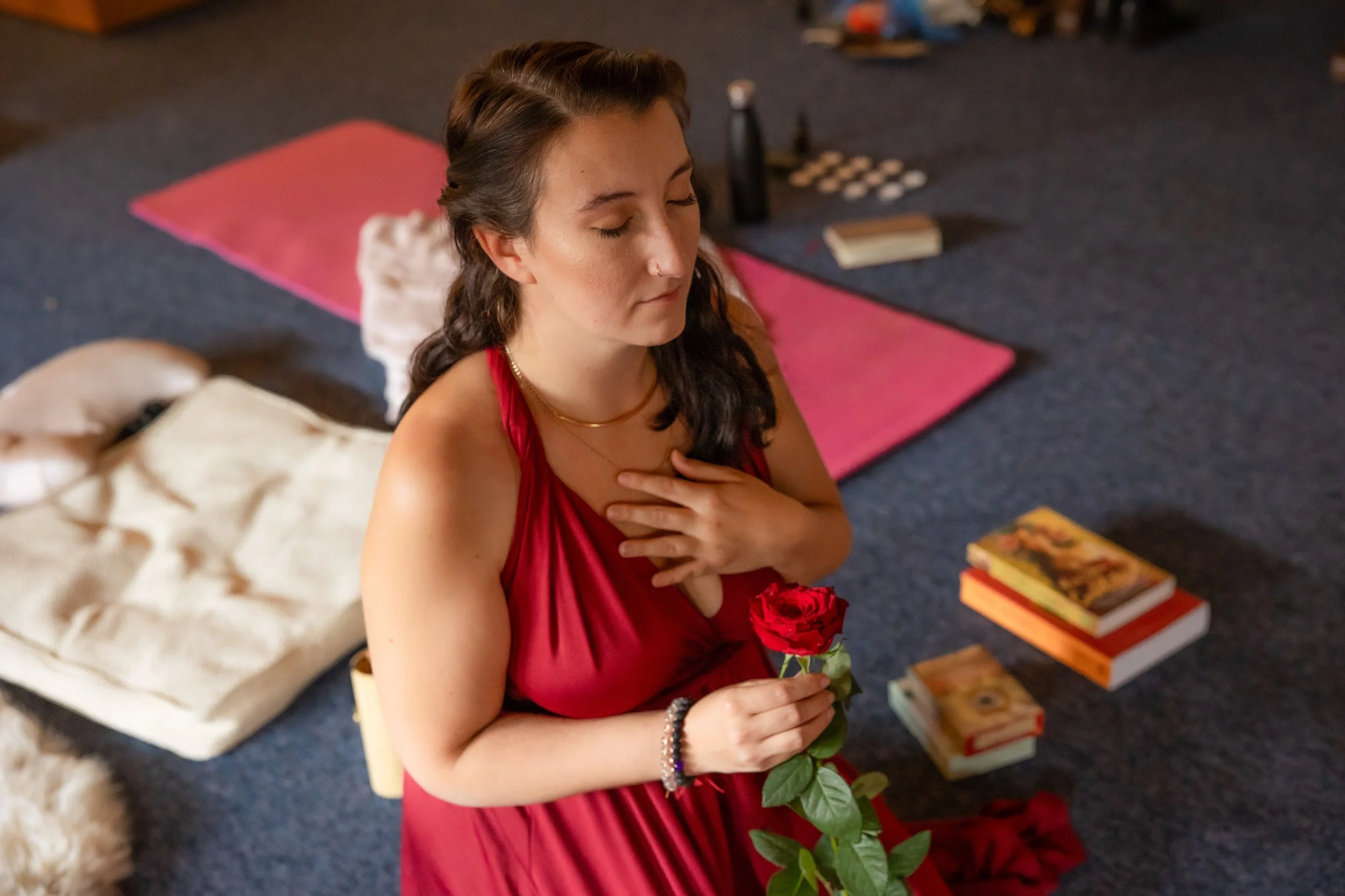 A woman in a red dress with closed eyes, holding a red rose close to her chest, in a room with books, a pink yoga mat, and other items on the floor.